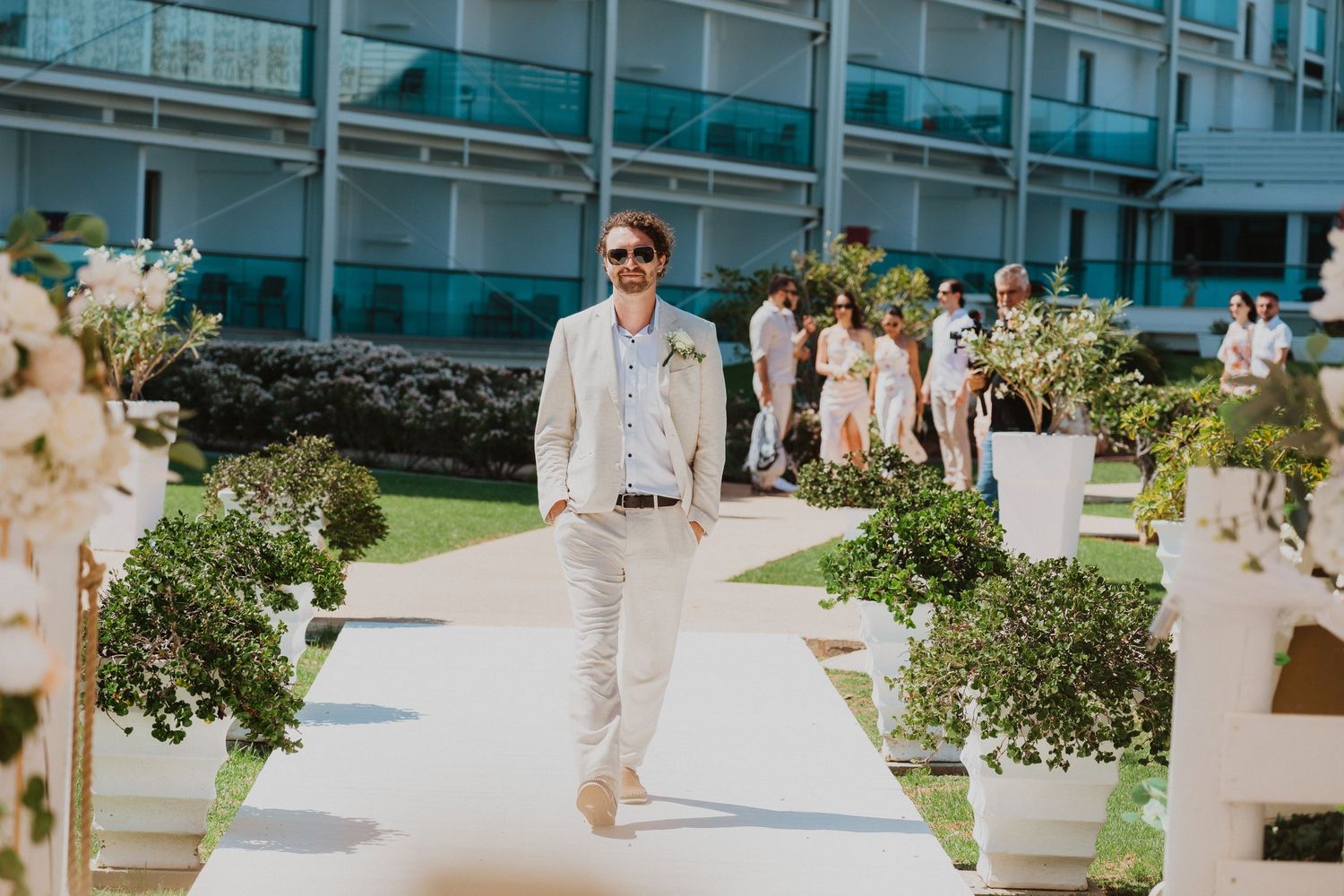 Beach wedding ceremony set up with white floral decor and an elegantly dressed individual walking down outdoor aisle.