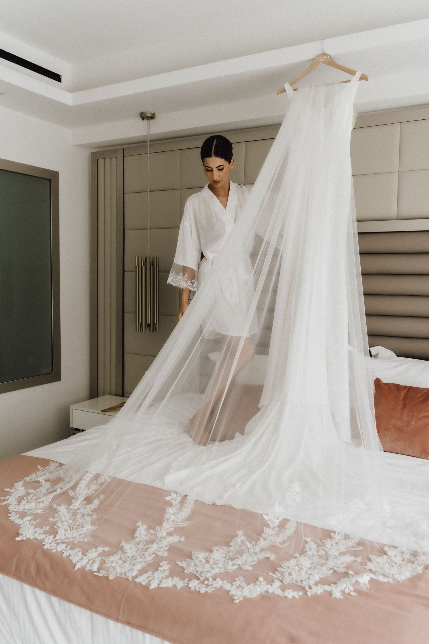 A flowing white wedding gown with lace details is displayed on a bed with blush linens.