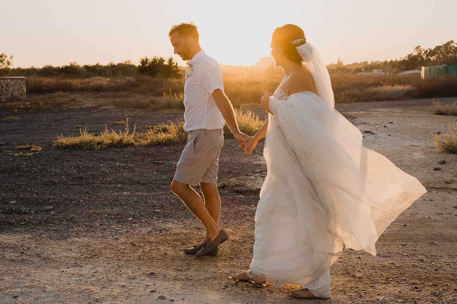 Desert wedding couple walking into golden sunset with flowing dress and casual attire.