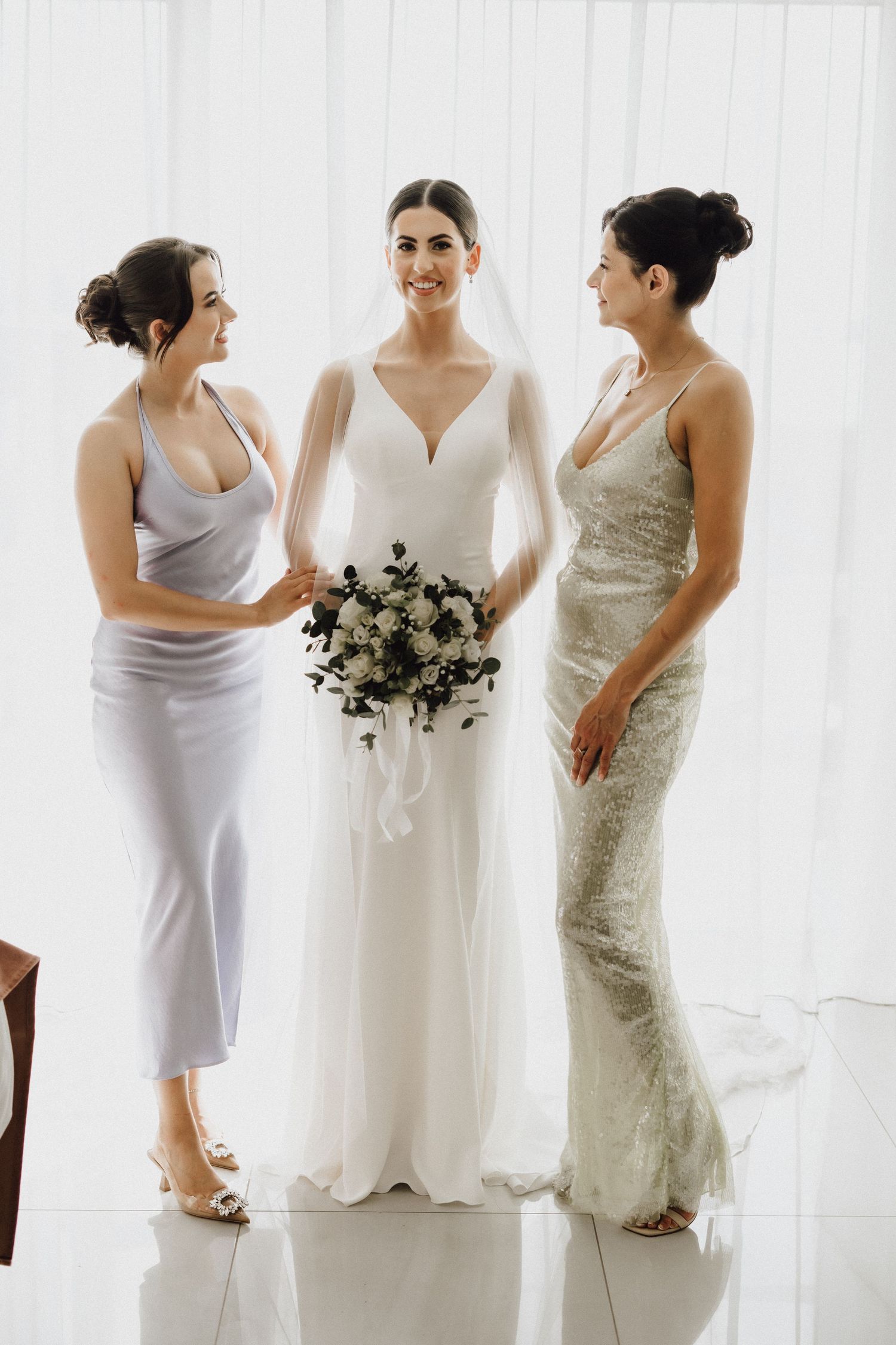 Three women pose together in elegant dresses, with the center figure wearing a white wedding gown and holding a bouquet.