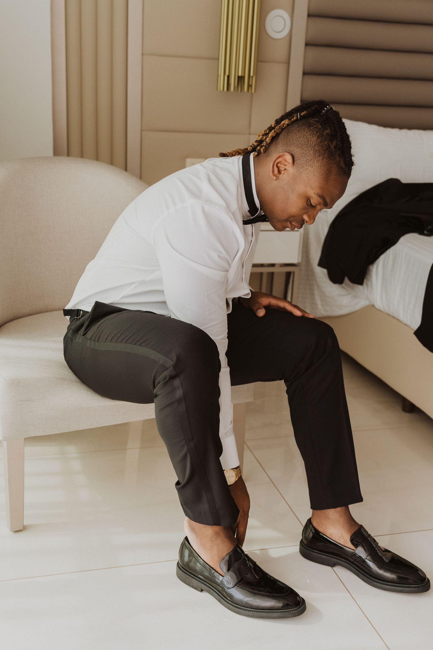 Person in formal attire adjusting their shoes while sitting on white furniture.