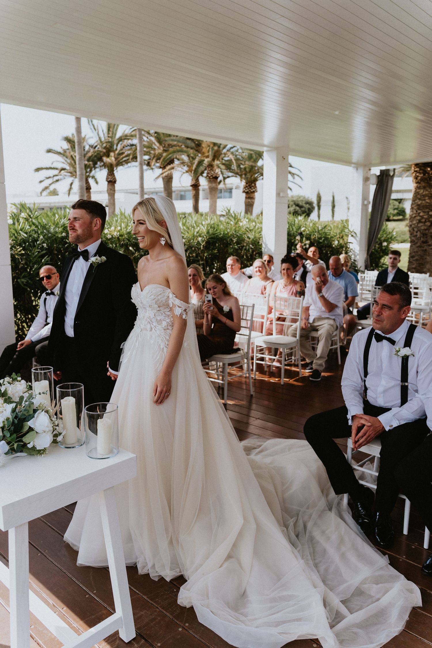 Elegant outdoor wedding ceremony under a covered patio with palm trees and white chairs for guests.