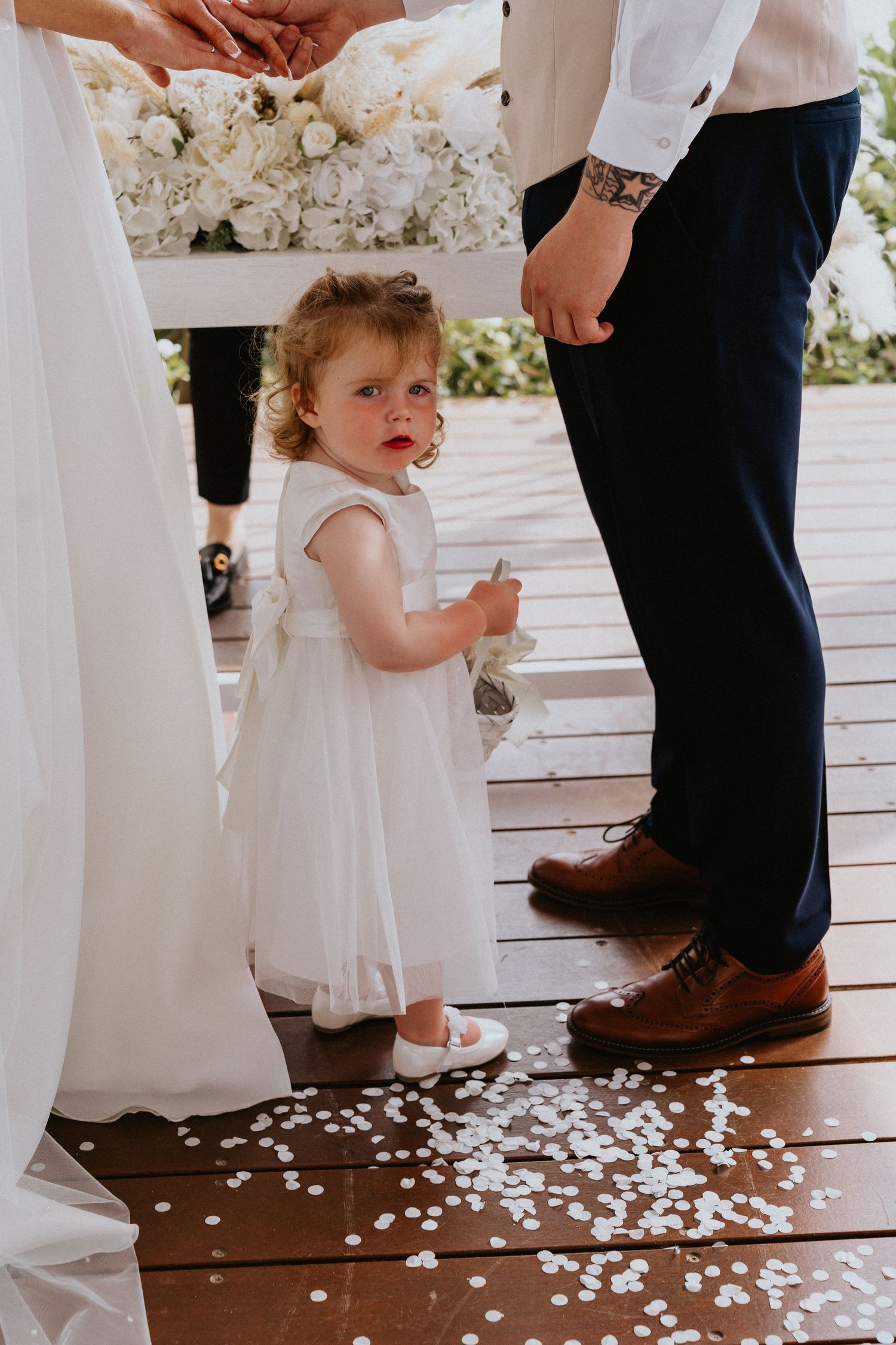 A young flower girl in white dress stands on wooden deck covered in confetti during wedding ceremony.