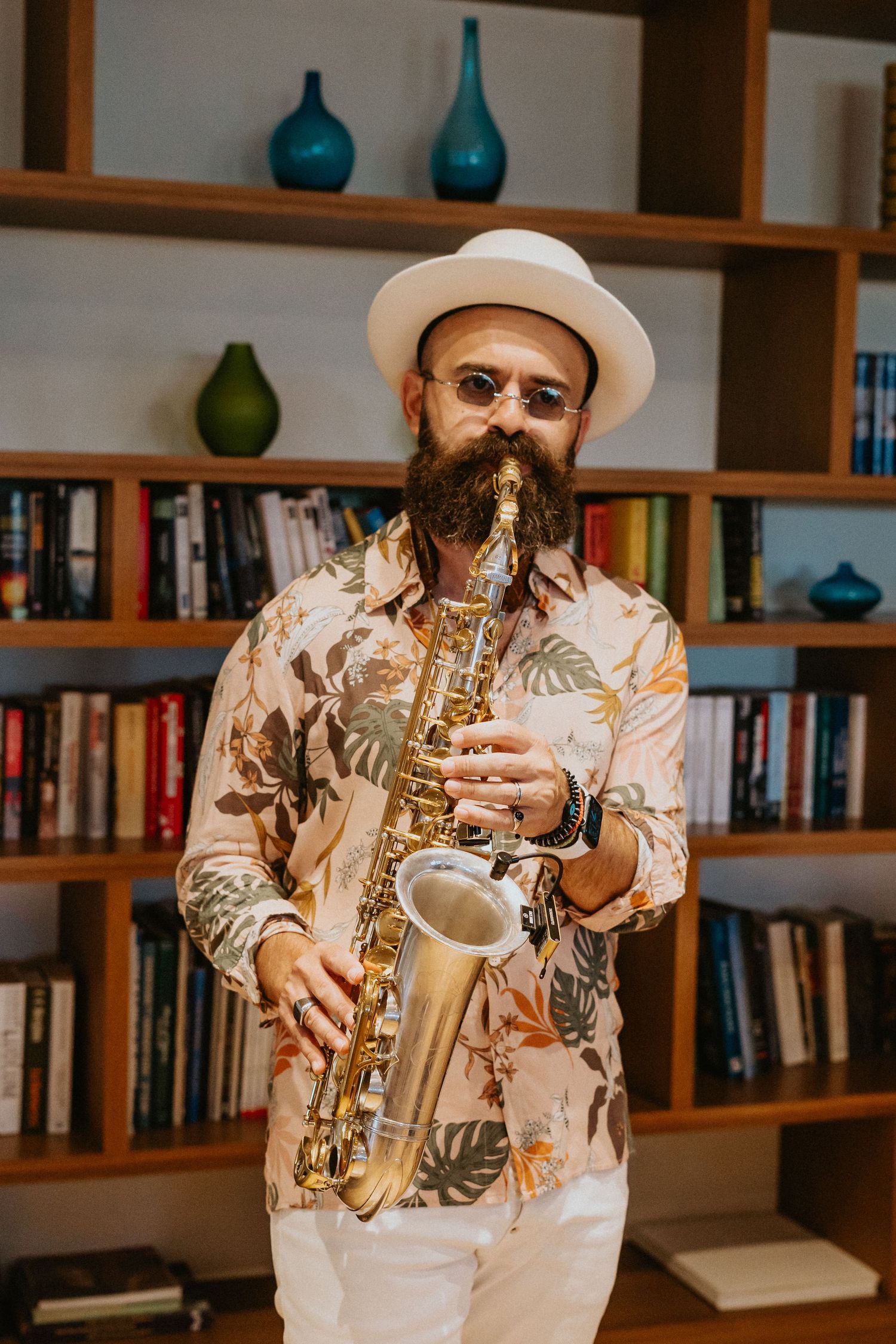 Musician plays saxophone in front of library bookshelf with turquoise vases while wearing floral shirt and white hat.