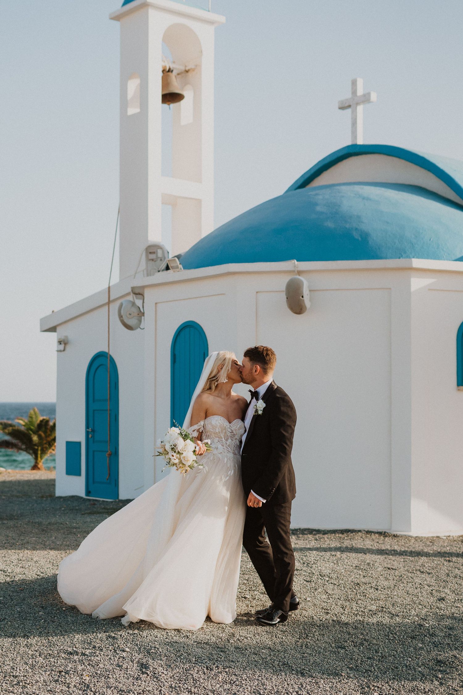 A romantic wedding photo in front of a white Mediterranean church with a blue dome and bell tower by the sea.