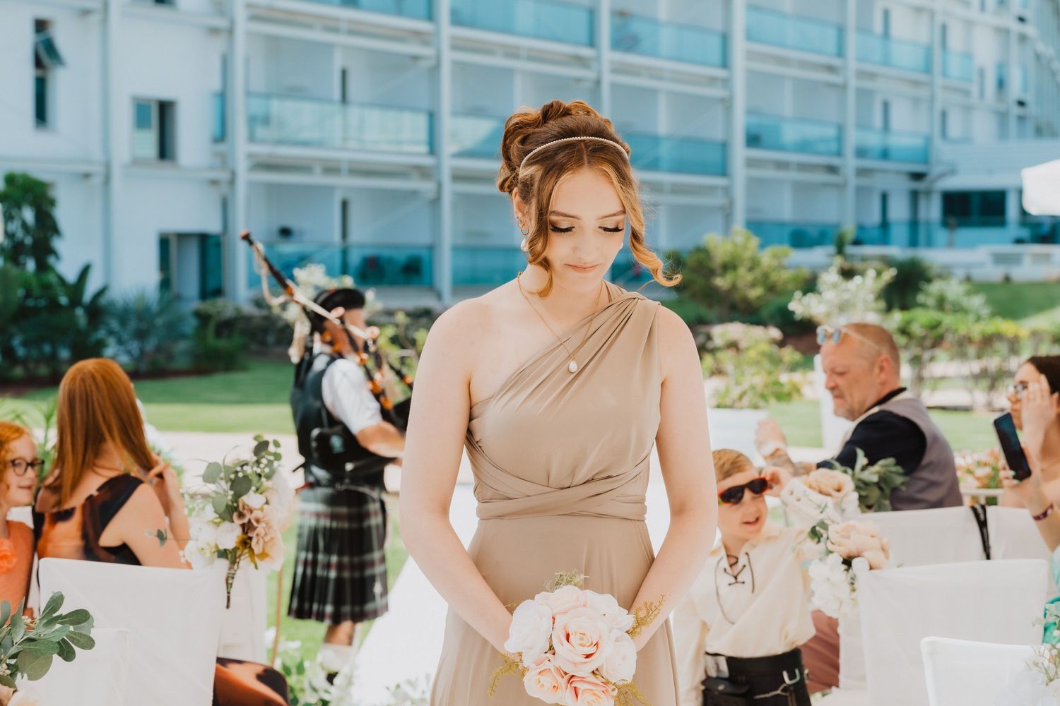 Wedding ceremony guests seated in white chairs with bridesmaids in beige dresses holding bouquets.
