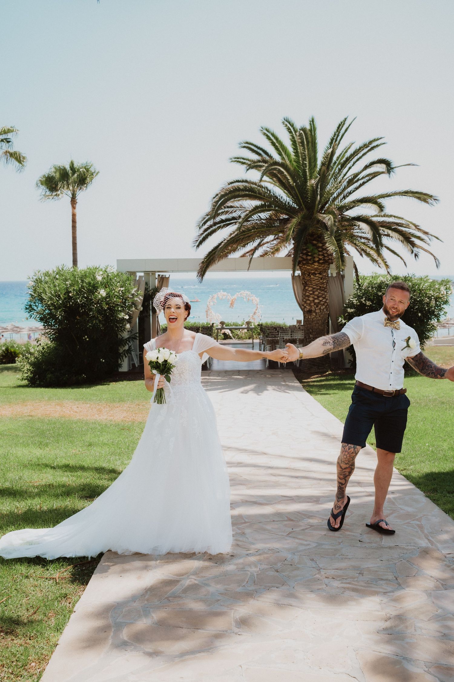 Wedding couple poses playfully on a beachfront walkway with palm trees in the background.