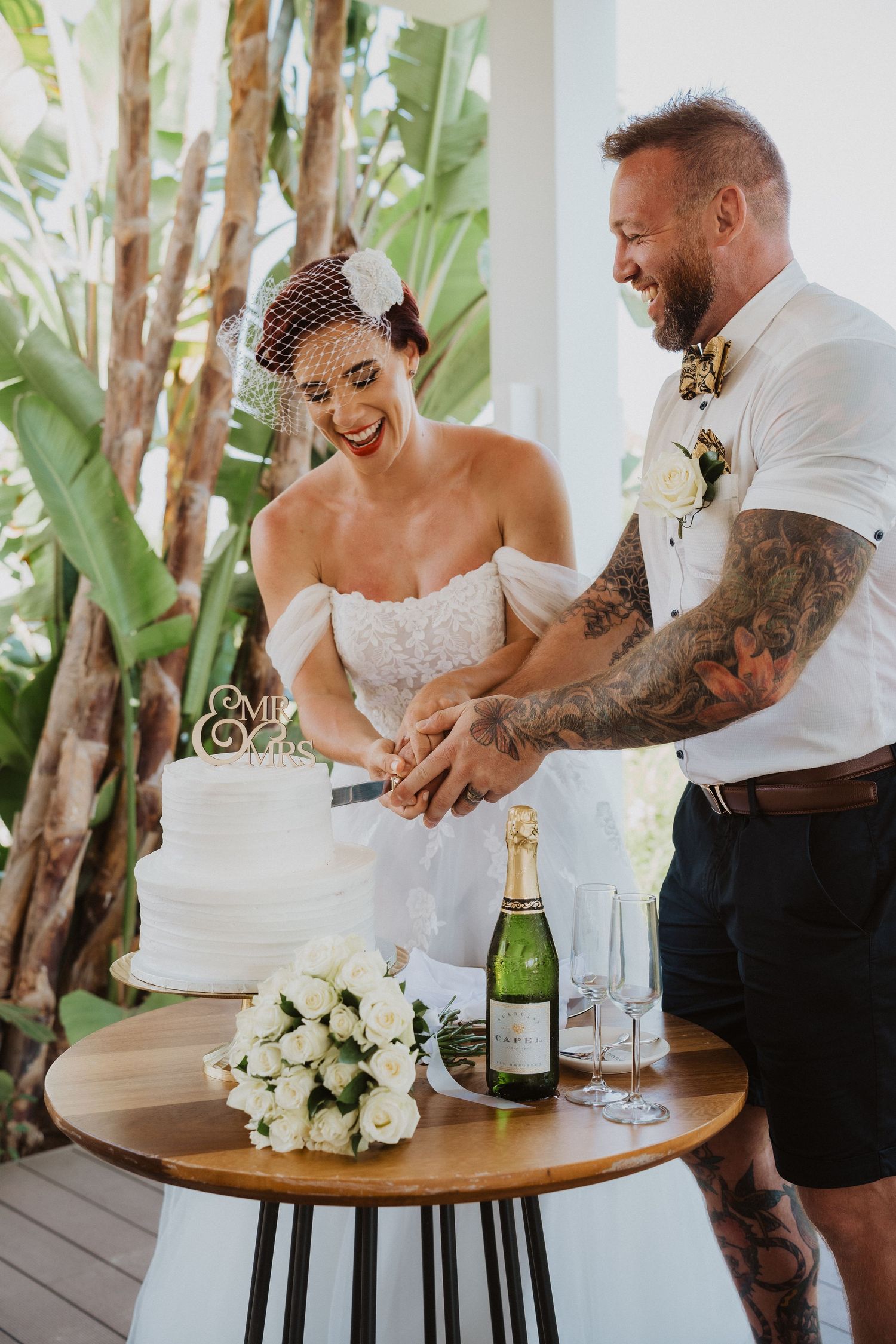 Happy couple cutting their white wedding cake together on outdoor patio.