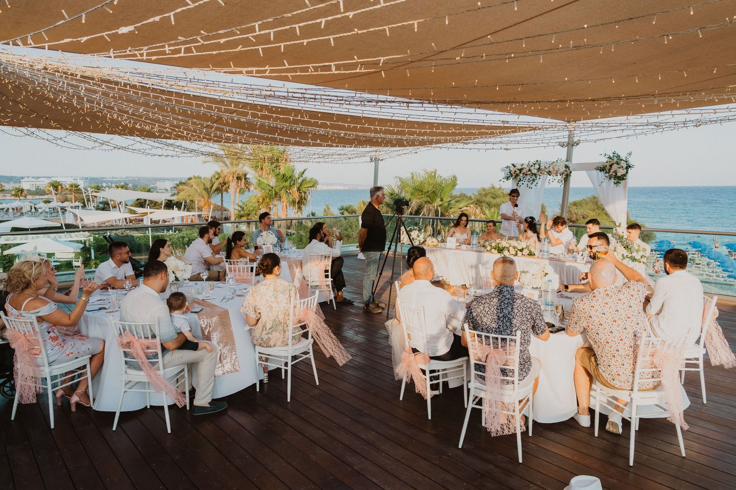 Wedding reception guests dine under string lights at an oceanfront venue with white tables and chairs.