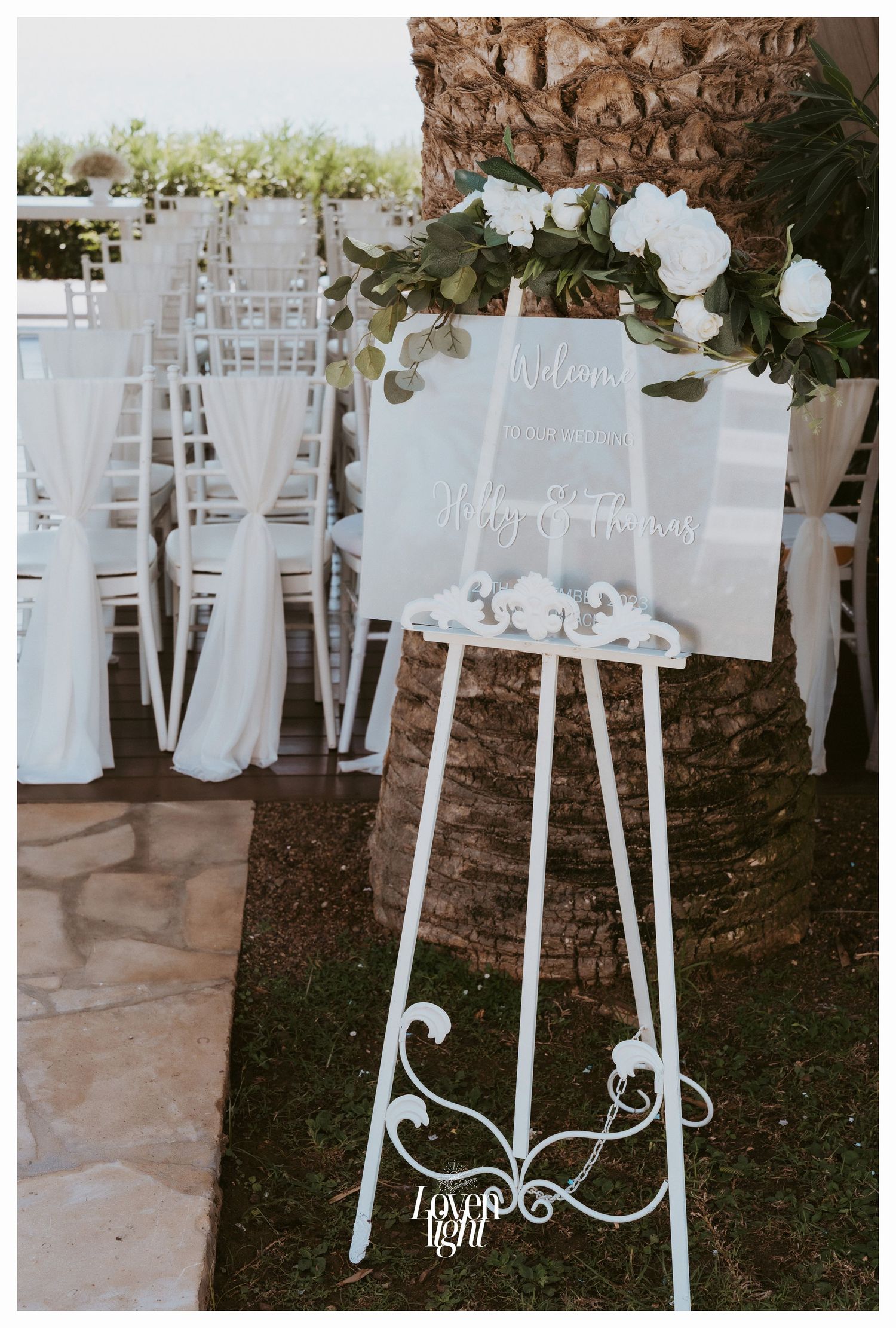 White welcome sign with floral decorations displayed on an elegant easel at a wedding ceremony.
