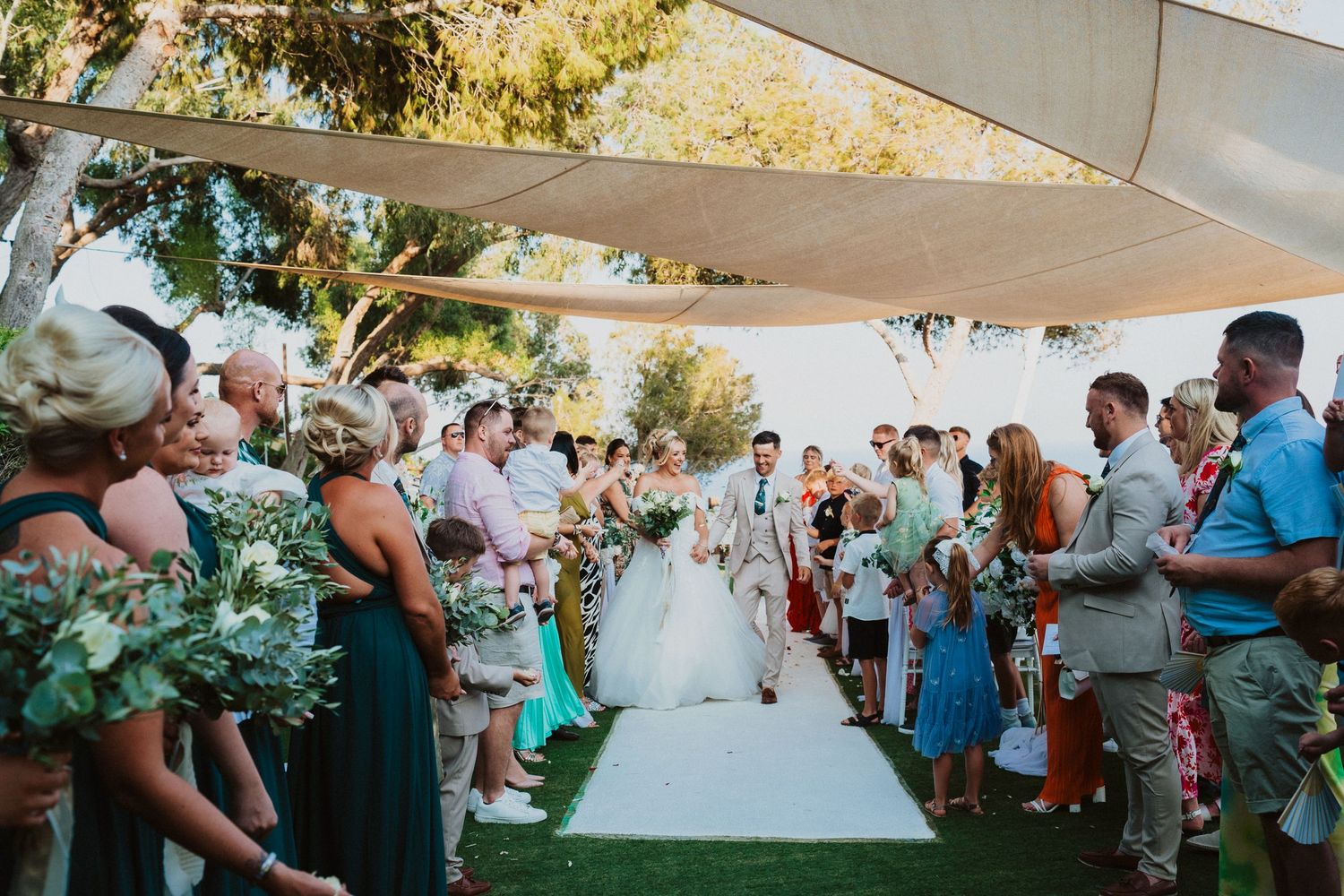 Wedding guests line a white aisle runner under an outdoor canopy during a beautiful garden ceremony.