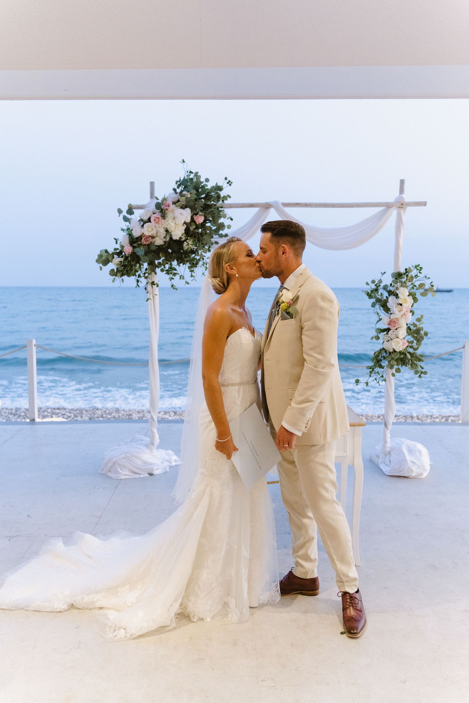 A romantic beach wedding ceremony under a floral arch with white draping and ocean views.