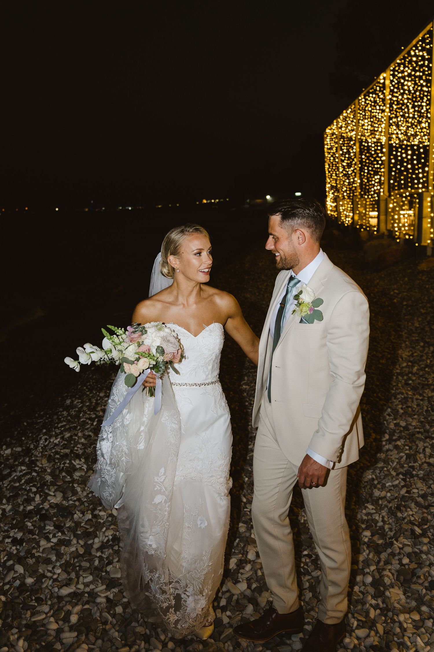 Wedding couple poses near illuminated structure on beach with bouquet at night.