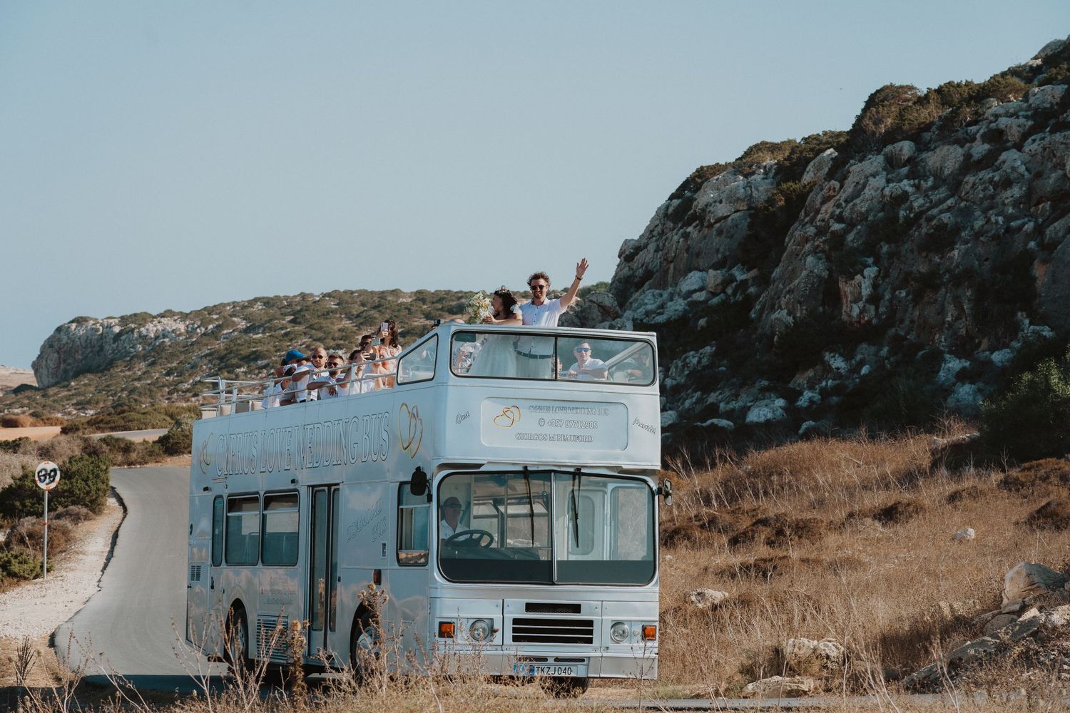 Double decker wedding bus driving along a coastal road with rocky cliffs in the background.