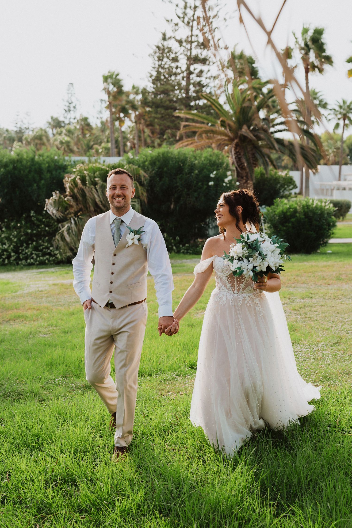 Newlywed couple holding hands while walking on green lawn with palm trees and tropical landscaping in background.