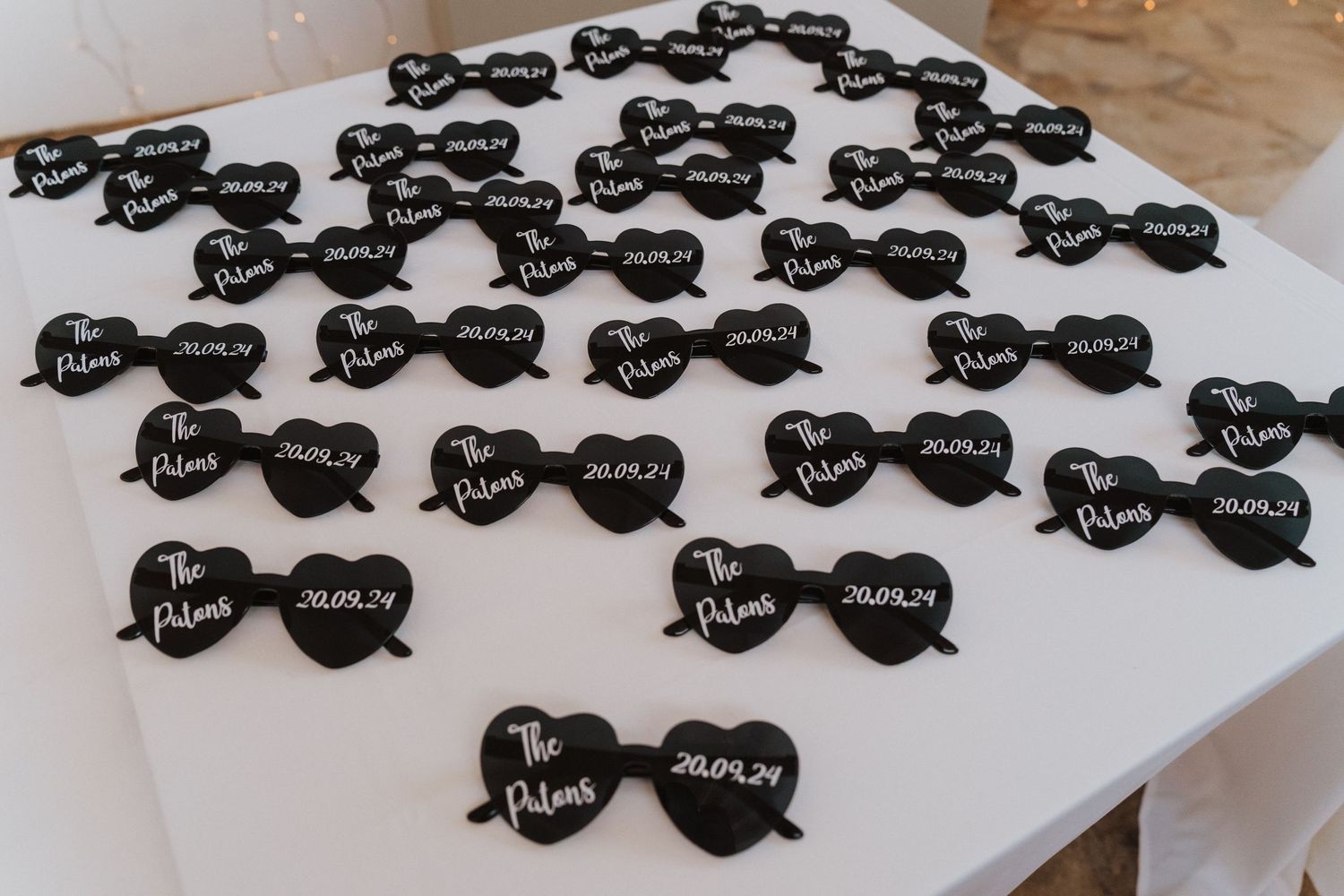 Black heart-shaped sunglasses with white text writing arranged neatly on a white surface for wedding favors.
