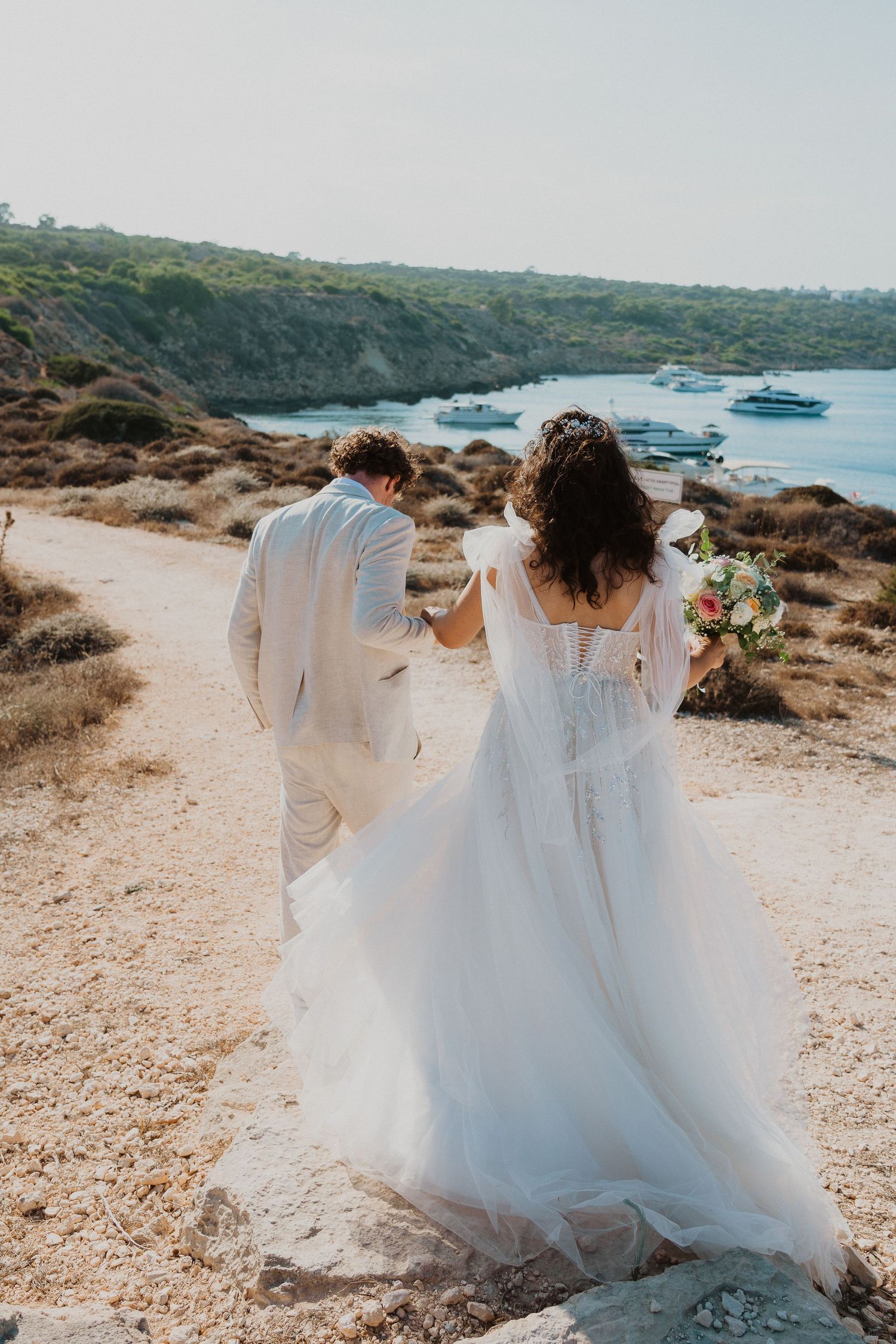 A couple in white wedding attire walk along a coastal path overlooking boats in a scenic Mediterranean bay.