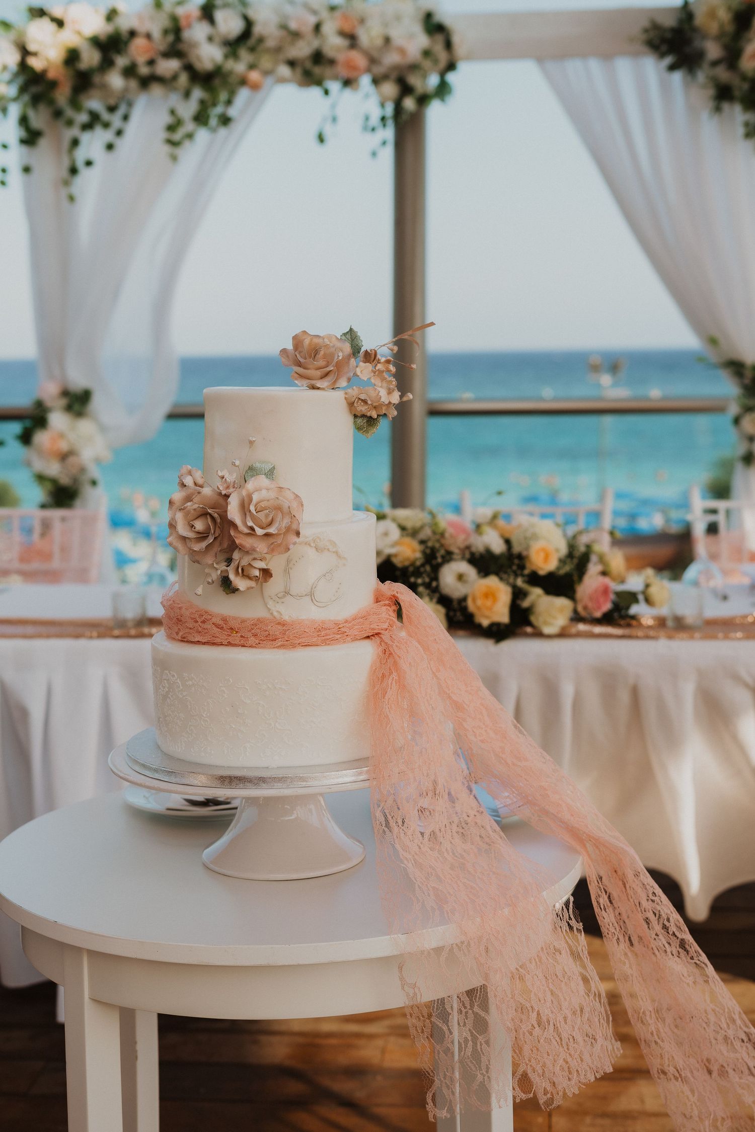 Elegant three-tiered white wedding cake with peach roses and lace on display at beachfront reception under draped gazebo.