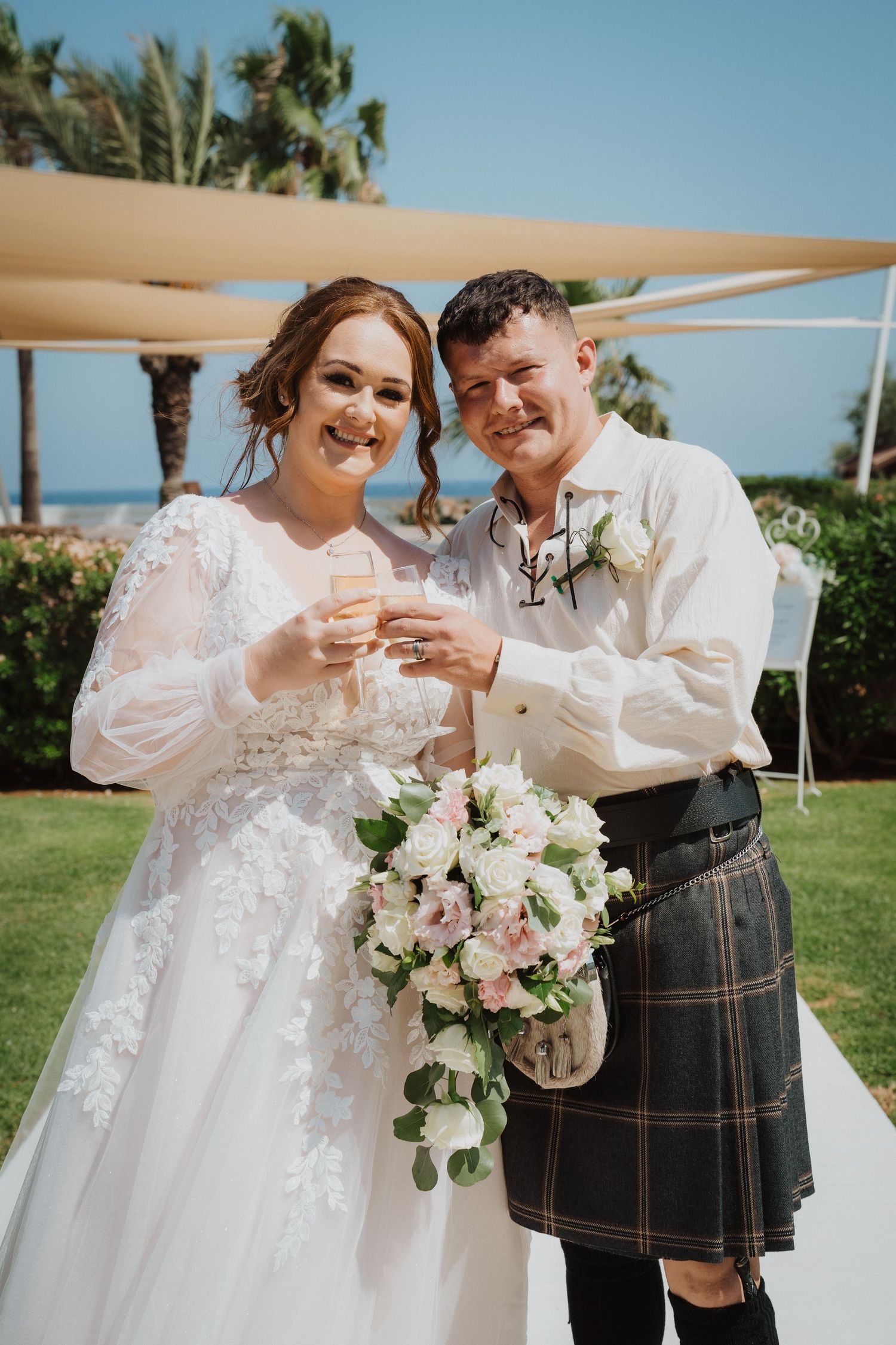 Traditional Scottish wedding couple sharing a celebratory moment with white floral bouquet under beige canopy.