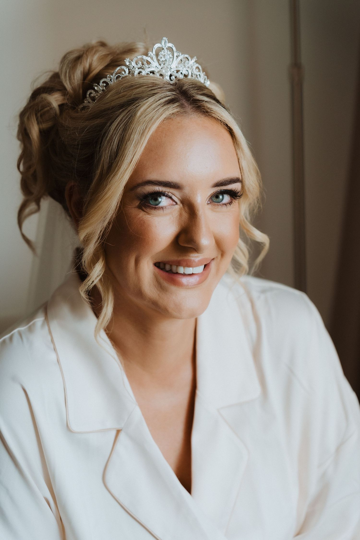 A bride in a white robe smiles while wearing a delicate crystal tiara and elegant bridal makeup.