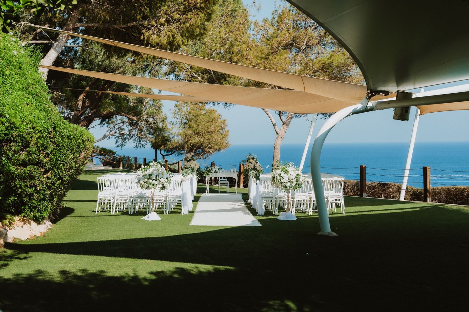 Elegant beach wedding venue with white chairs under tan shade sails and ocean views.