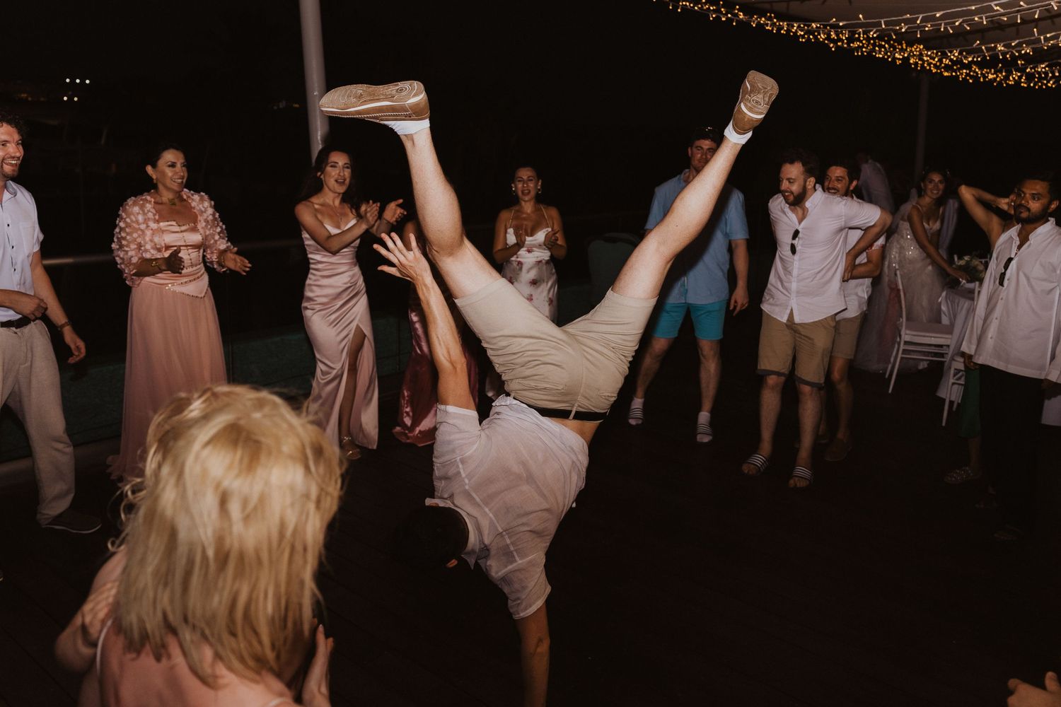 Wedding guest performs impressive dance moves on reception dance floor under string lights.