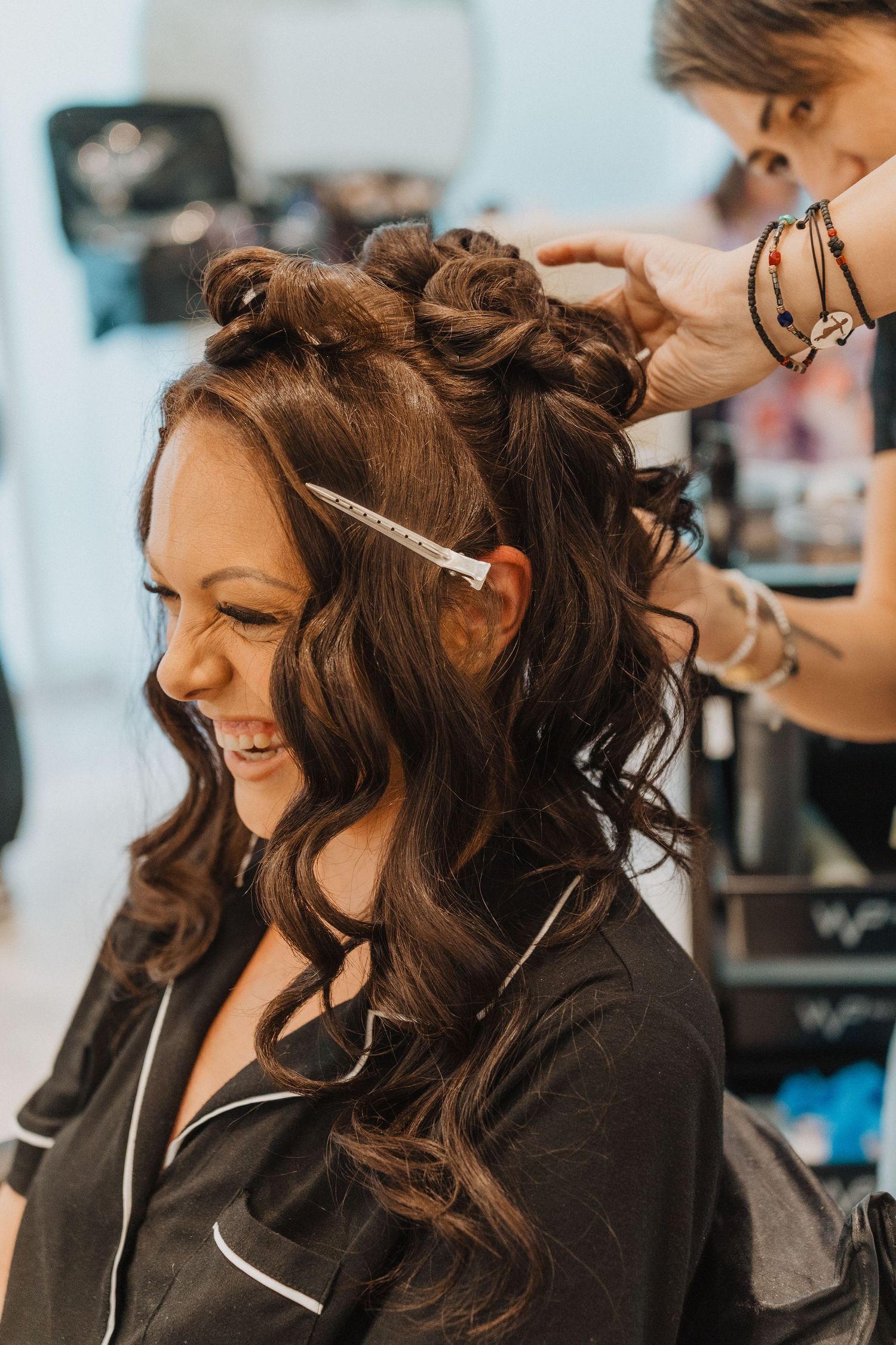 Stylist works on creating an elegant updo hairstyle with loose curls while client smiles in salon chair.