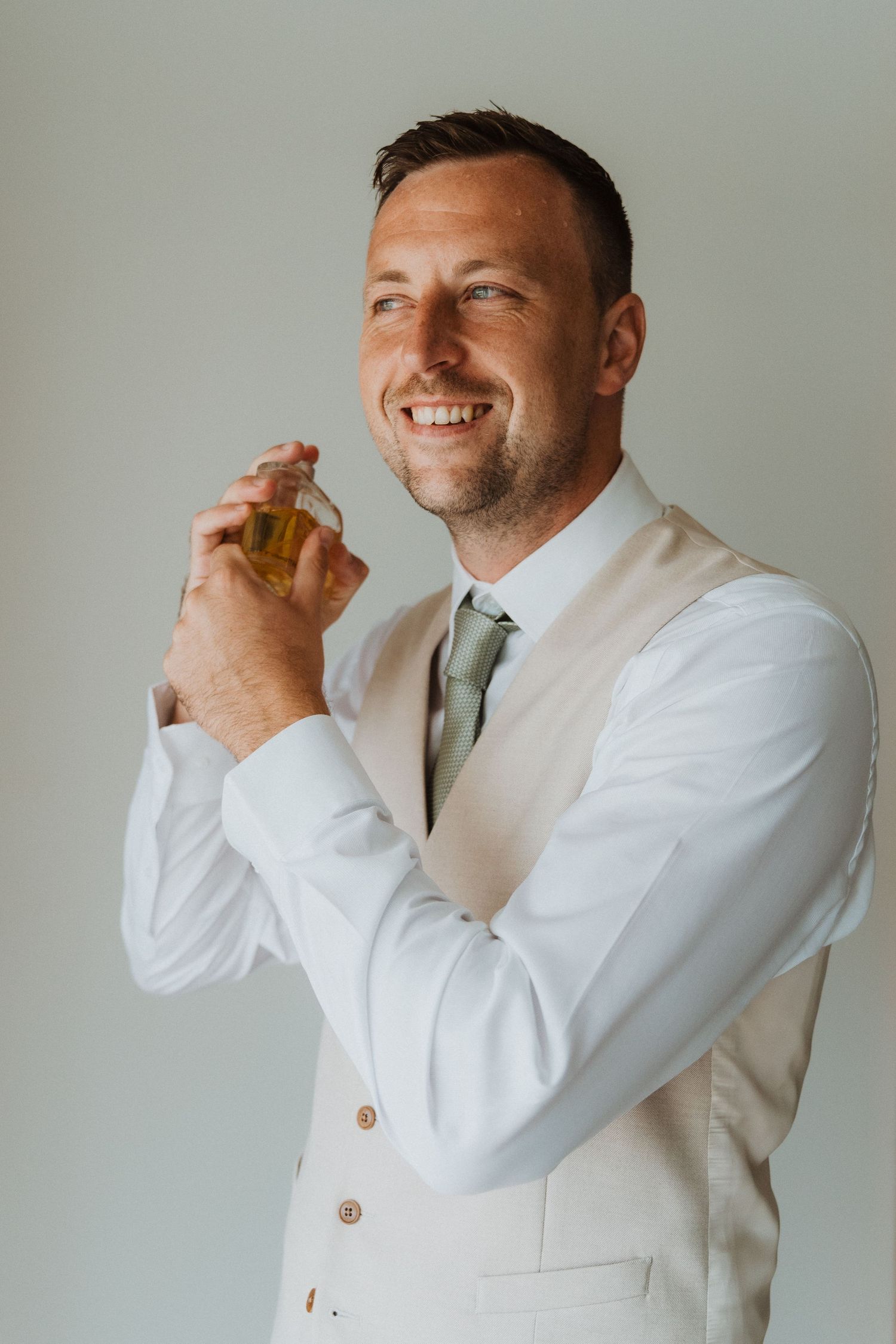 A person in a white dress shirt and vest holding a small perfume bottle while smiling against a light background.