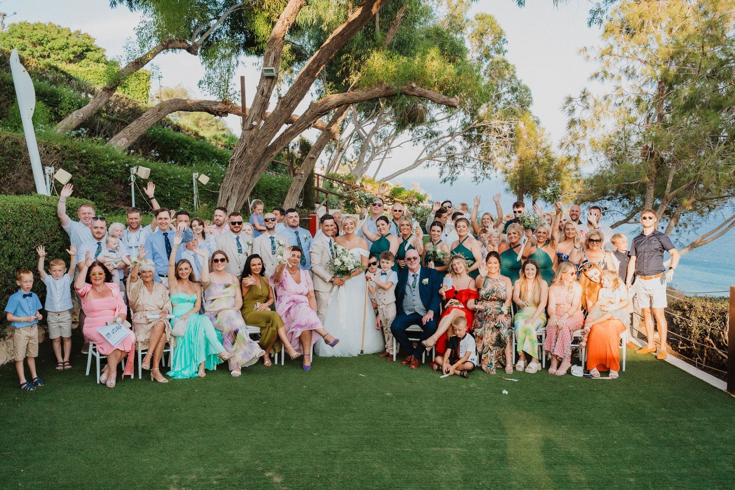 Large wedding party group photo on a grassy lawn with ocean view and trees in background.