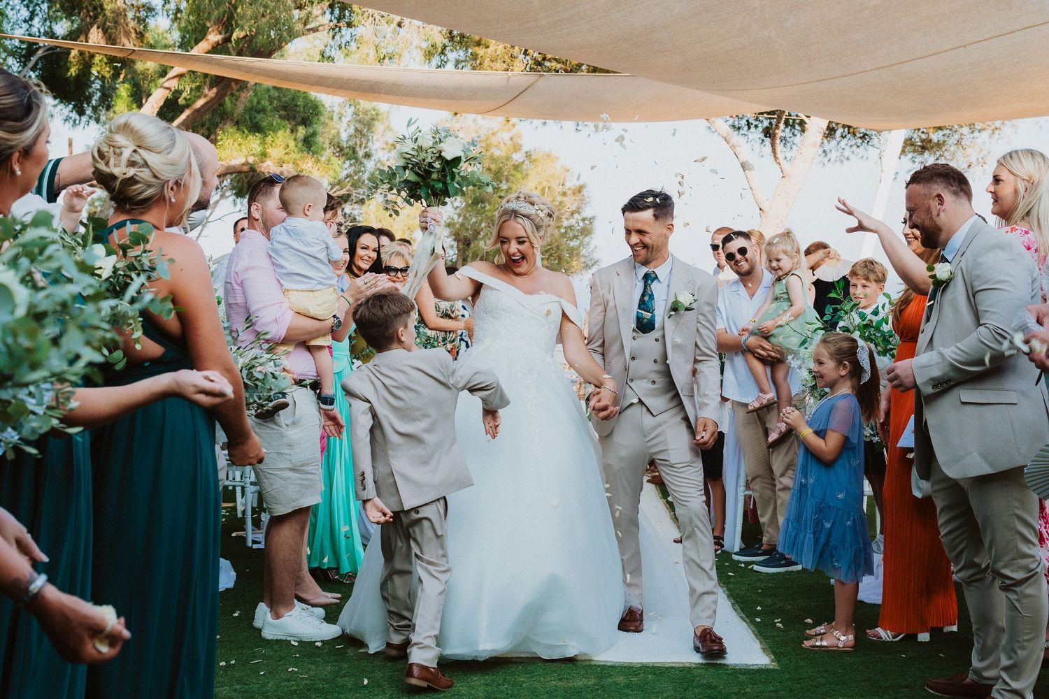 Wedding party celebrates during outdoor ceremony under white canopy with guests in green and blue attire.