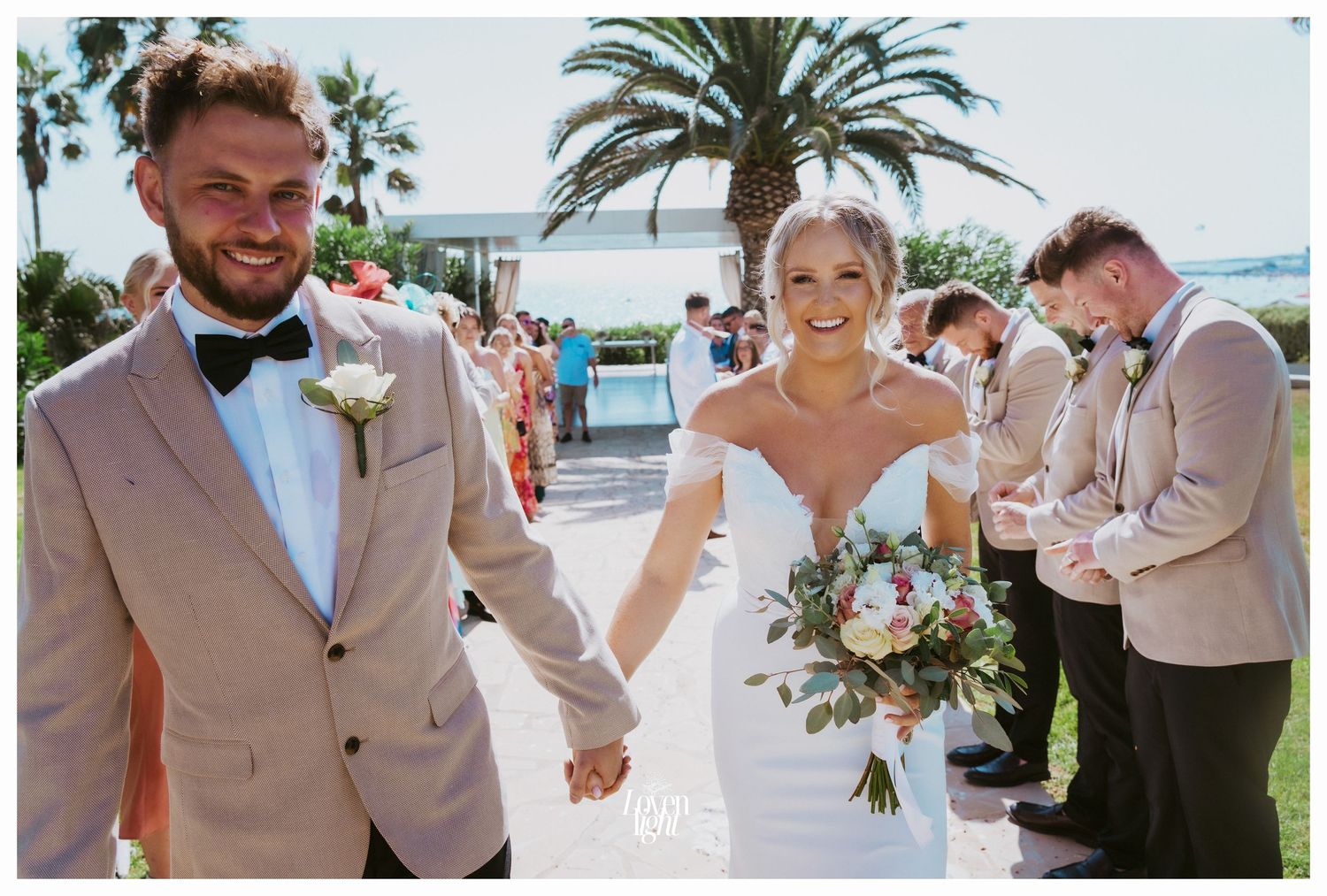 A couple celebrates joyfully during their wedding ceremony on a sunny beach with palm trees in the background.