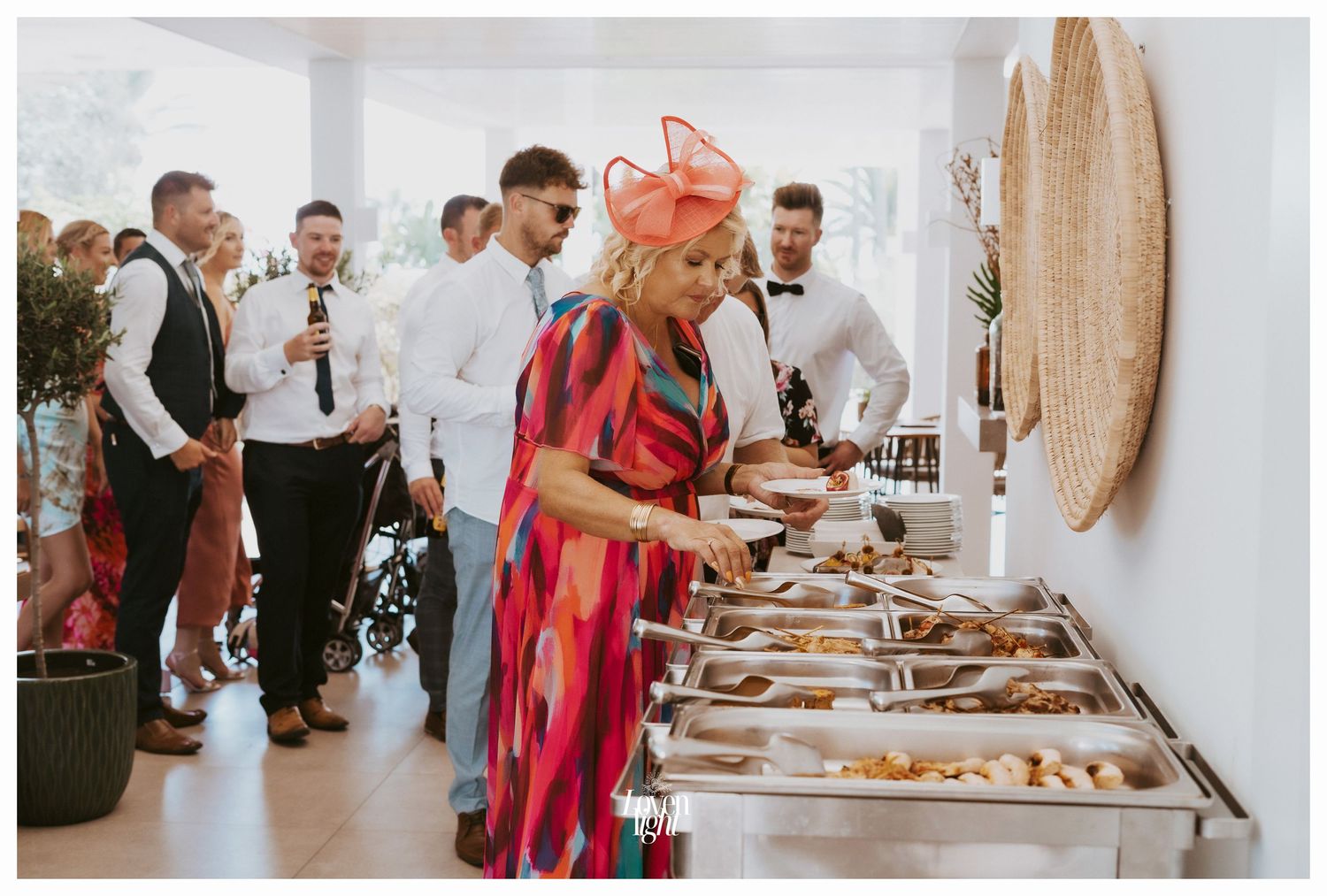Wedding guests serving themselves from an elegant buffet line with woven decor on the wall.