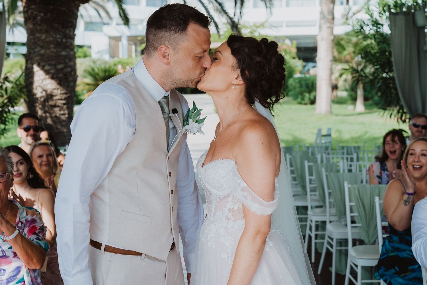 A romantic wedding ceremony kiss shared outdoors with tropical palm trees and white chairs in the background.