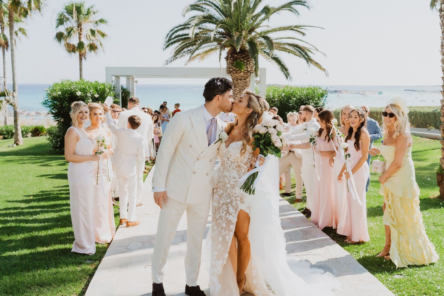 Wedding party in soft pink dresses and white suits gather for outdoor photos under palm trees near the beach.
