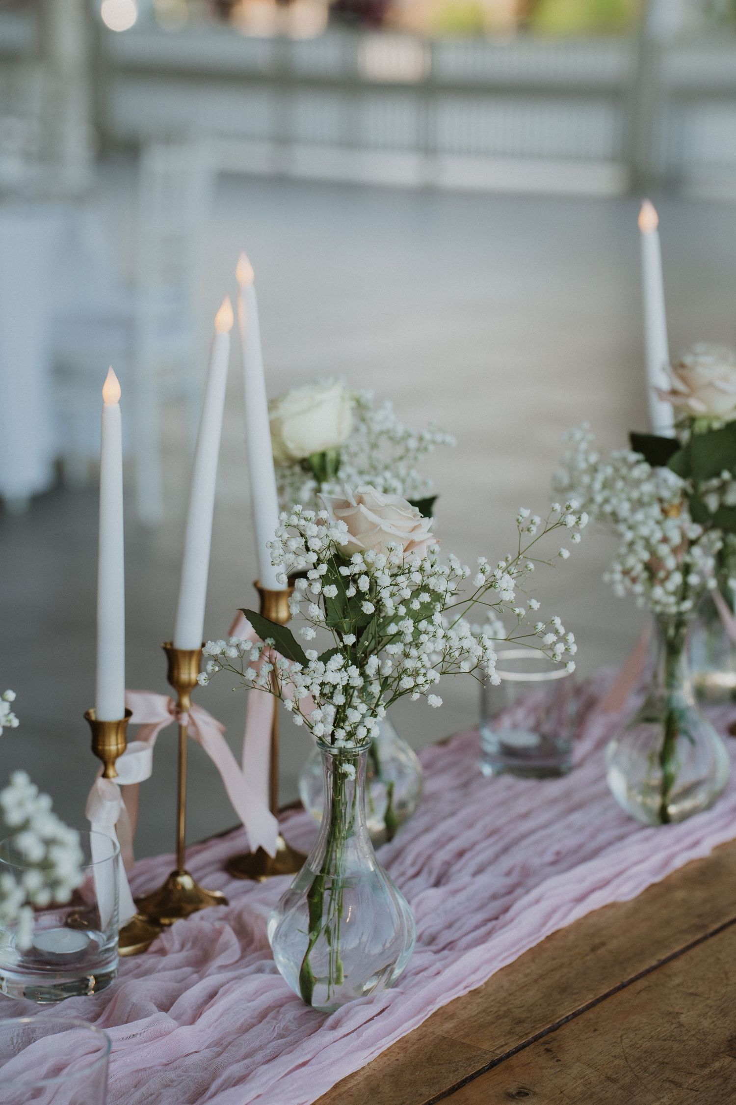 Elegant table setting with white taper candles, baby's breath flowers in glass vases, and pink fabric runner on wooden surface.