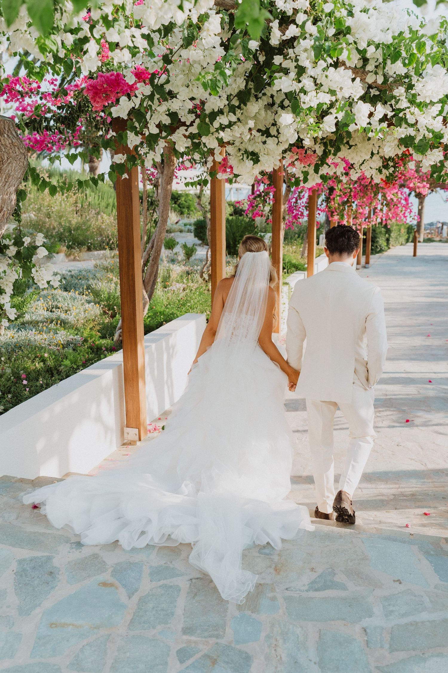 A couple walks under a floral pergola with pink and white blooms on a stone pathway at a destination wedding venue.