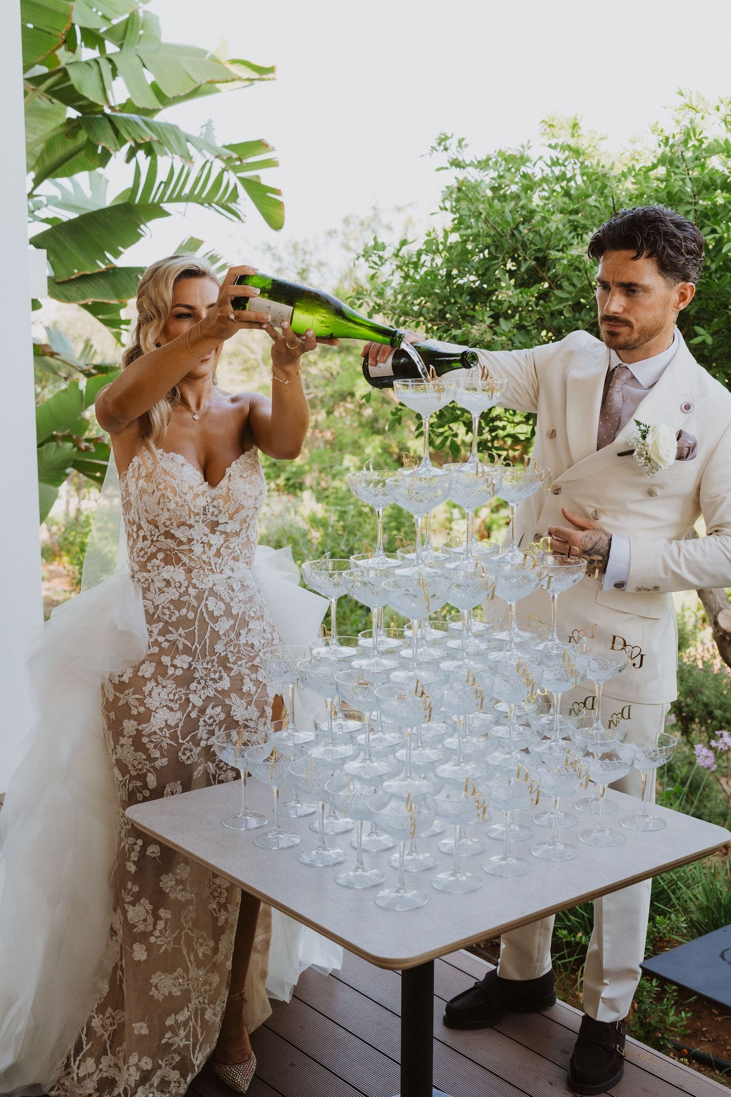 A festive champagne tower being carefully assembled on an outdoor patio with lush greenery in the background.