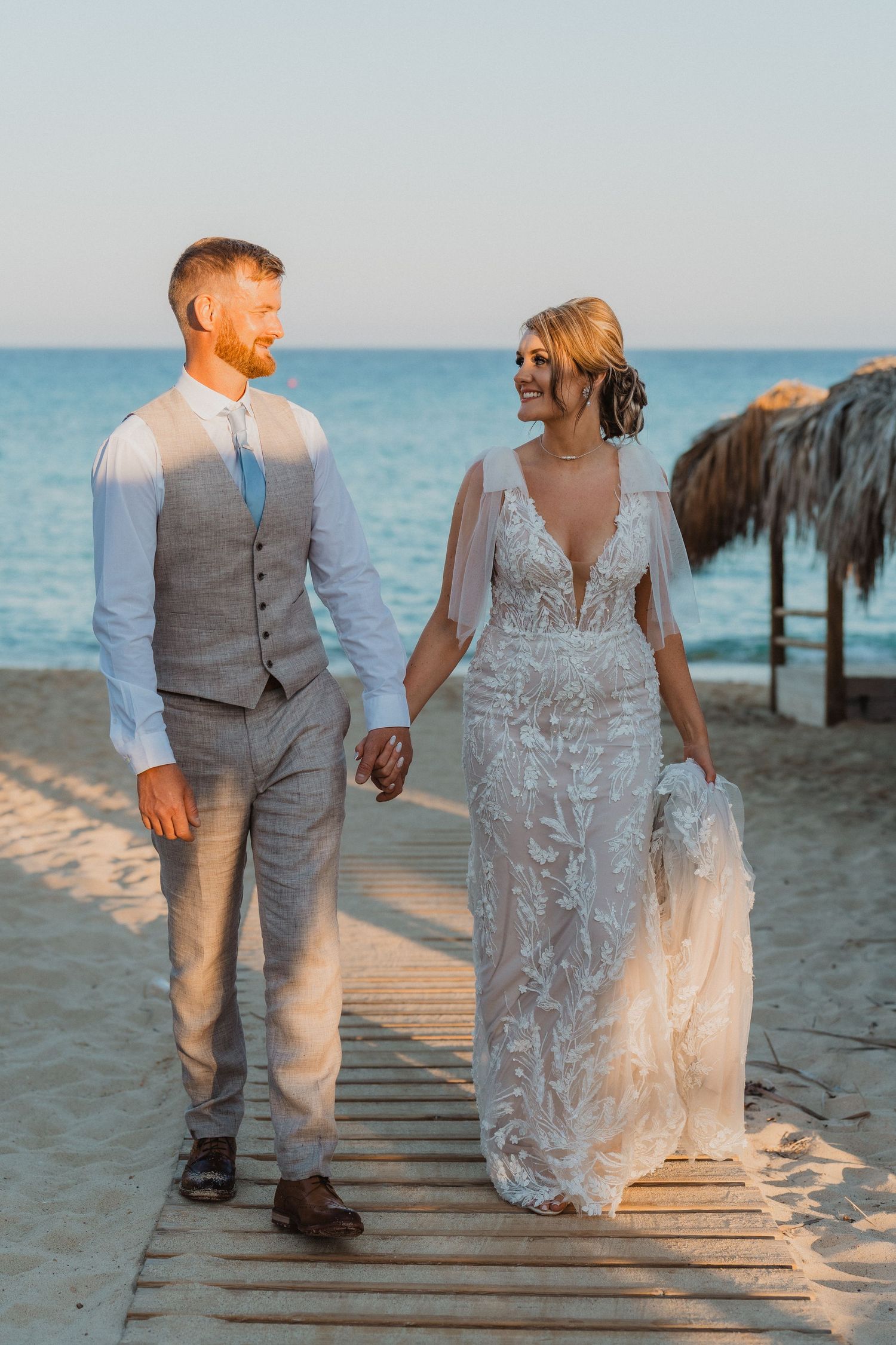 Couple in wedding attire walks along wooden boardwalk on tropical beach at sunset.