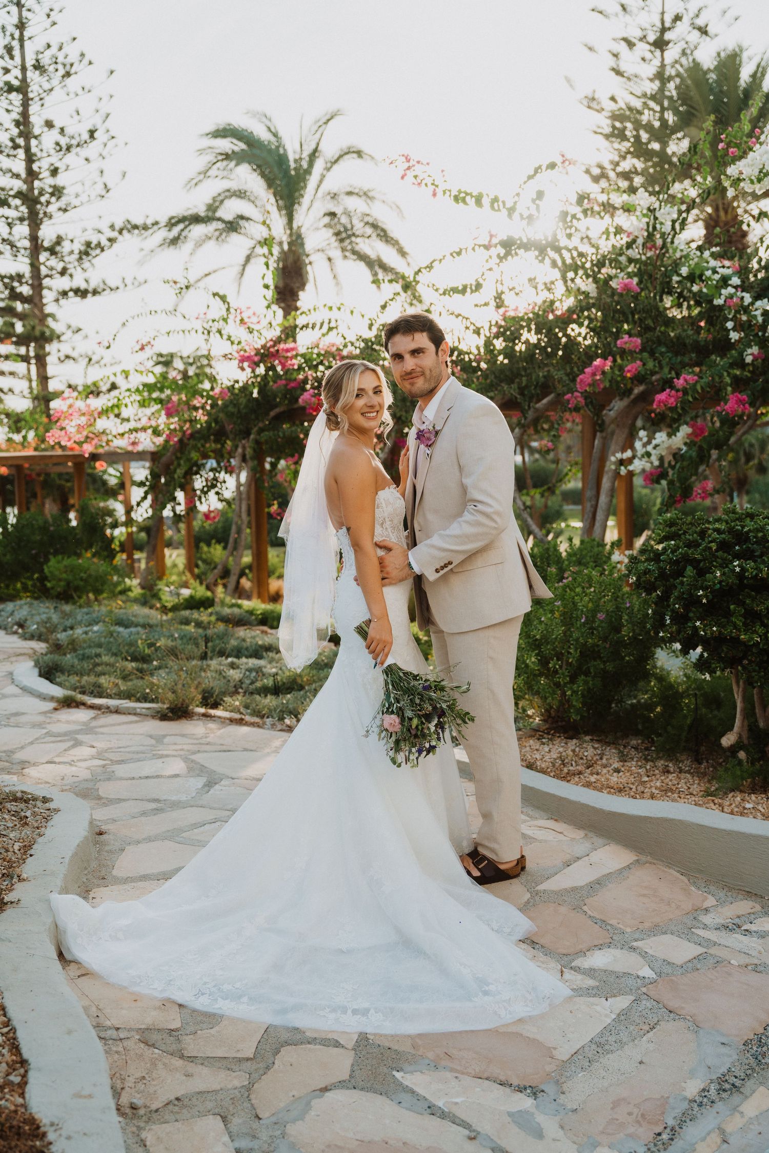 Joyful wedding couple poses on stone pathway surrounded by lush tropical landscaping.