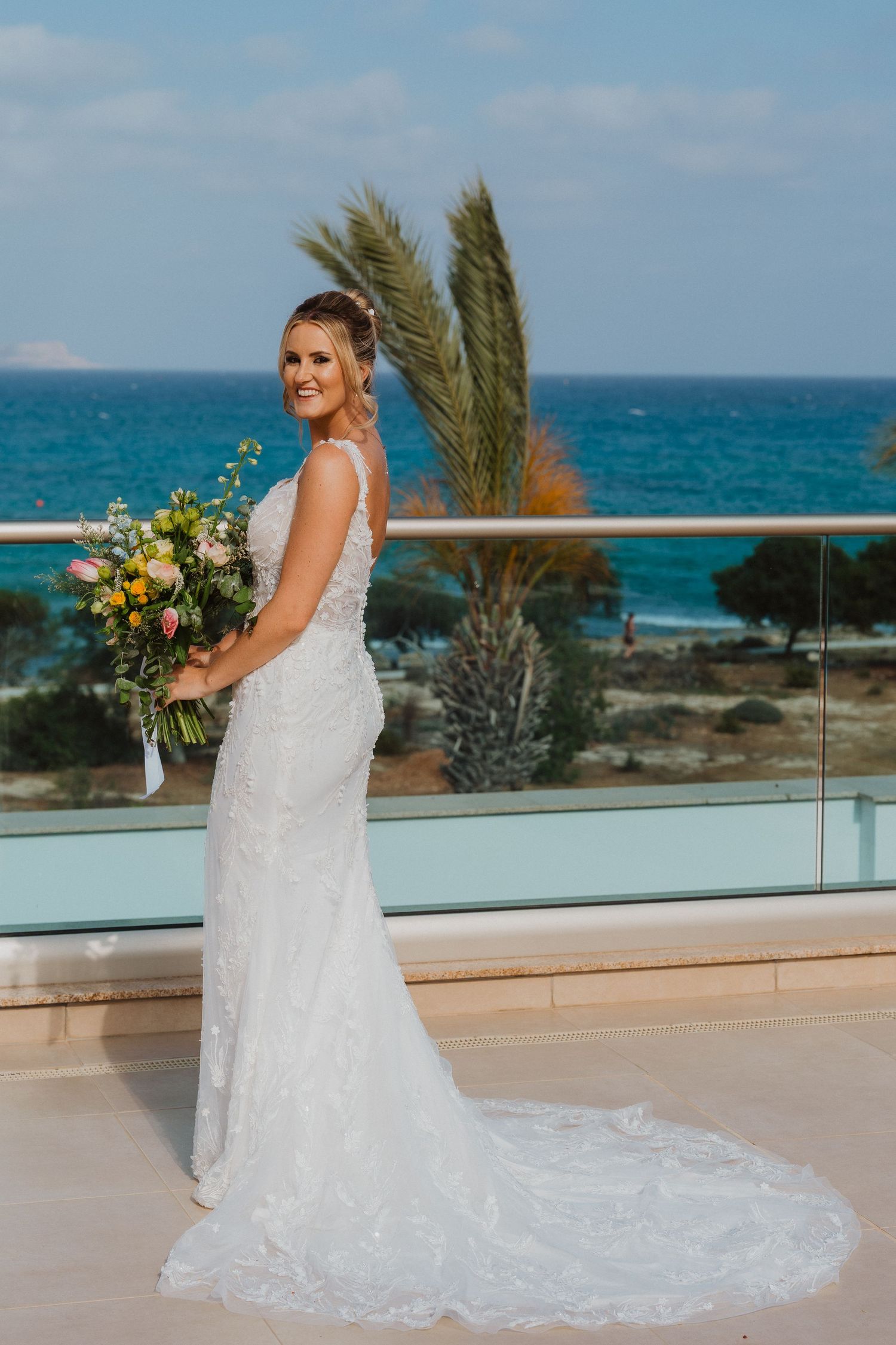 A bride in a white lace wedding gown with train poses on an oceanfront balcony holding a yellow and white bouquet.