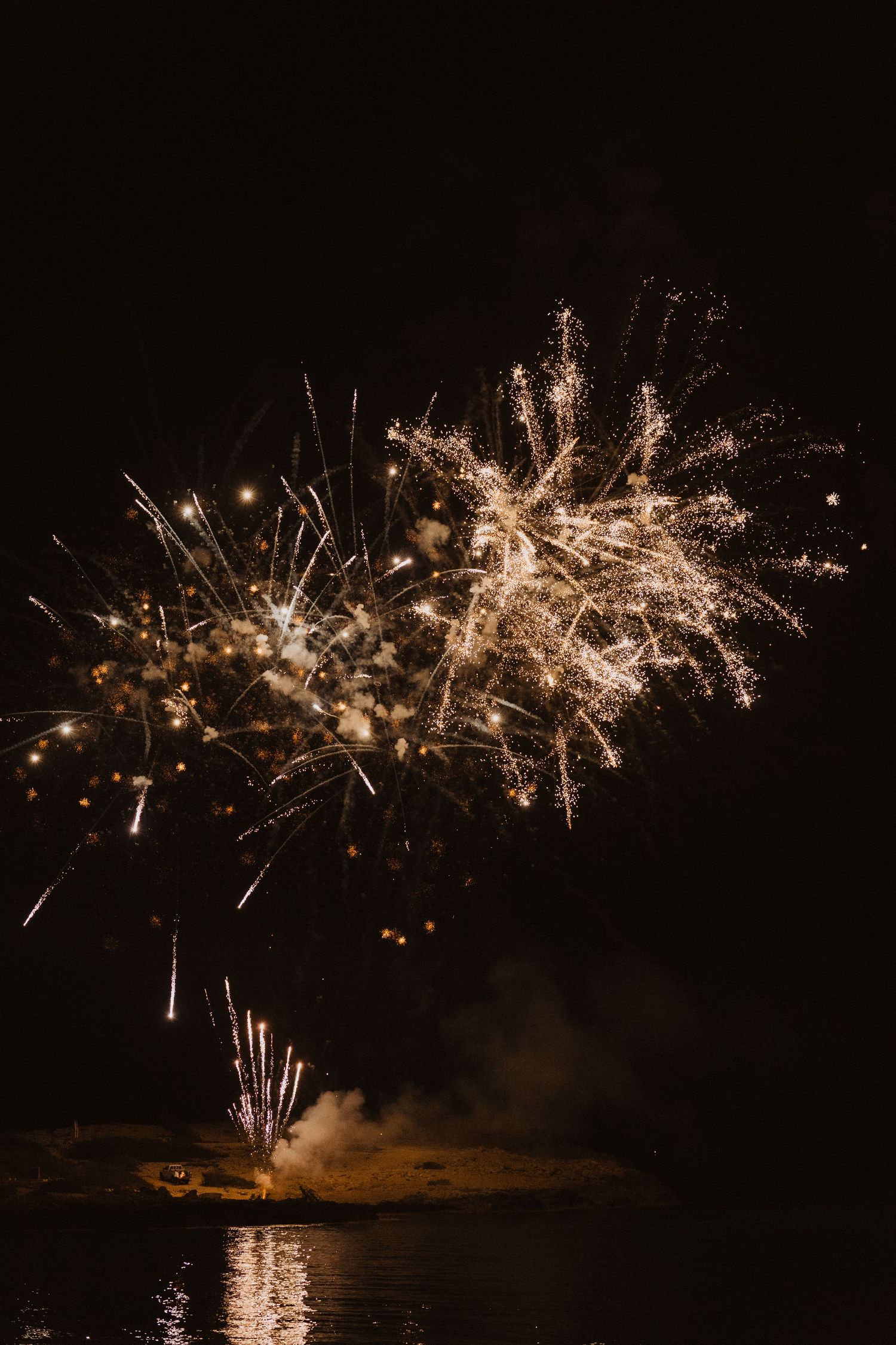 Spectacular fireworks display reflecting over water with golden sparks illuminating the night sky.