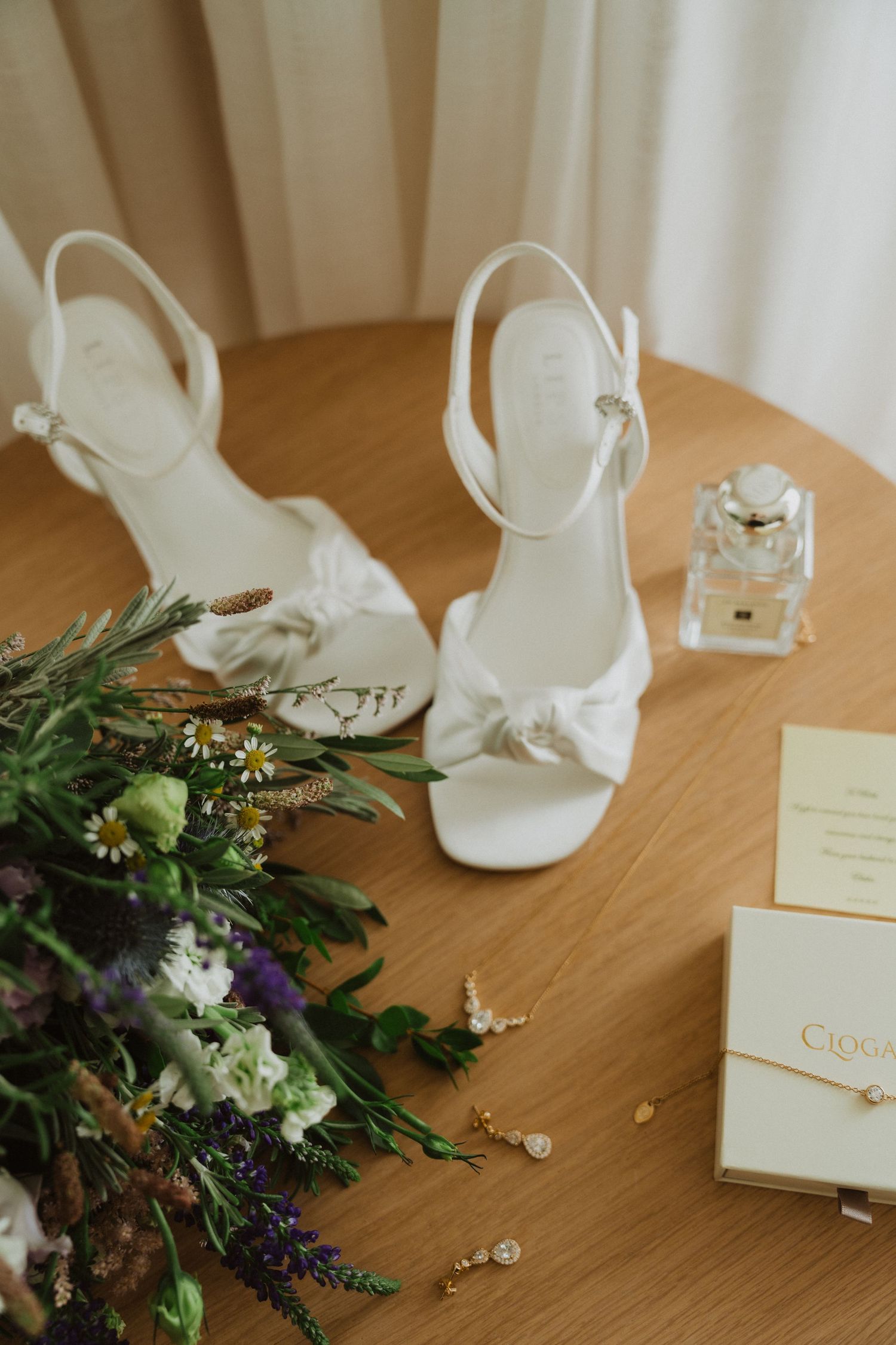 Wedding accessories including white heels, jewelry, and floral arrangements on a wooden surface.