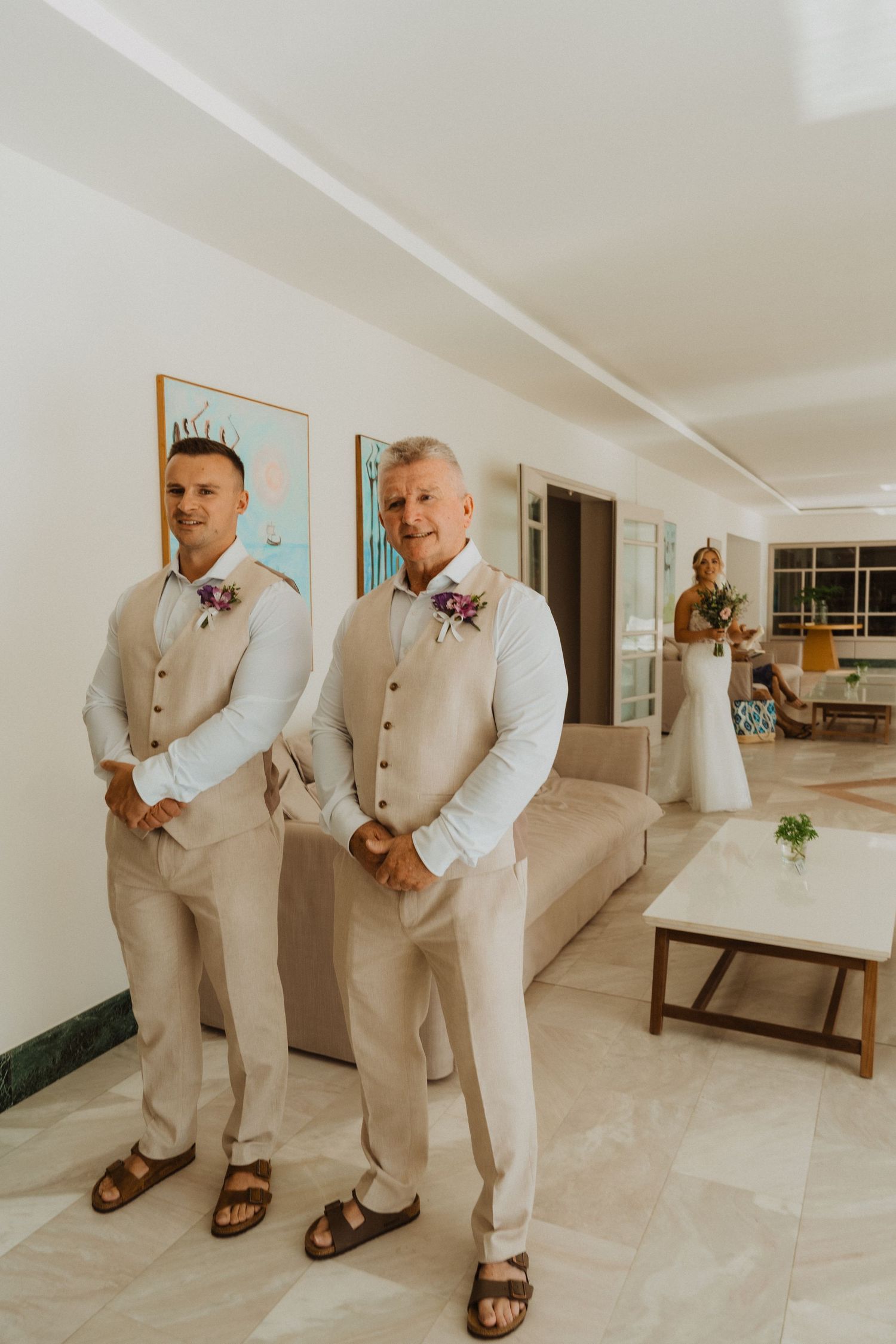 Wedding party members in beige suits pose playfully on white marble floors.