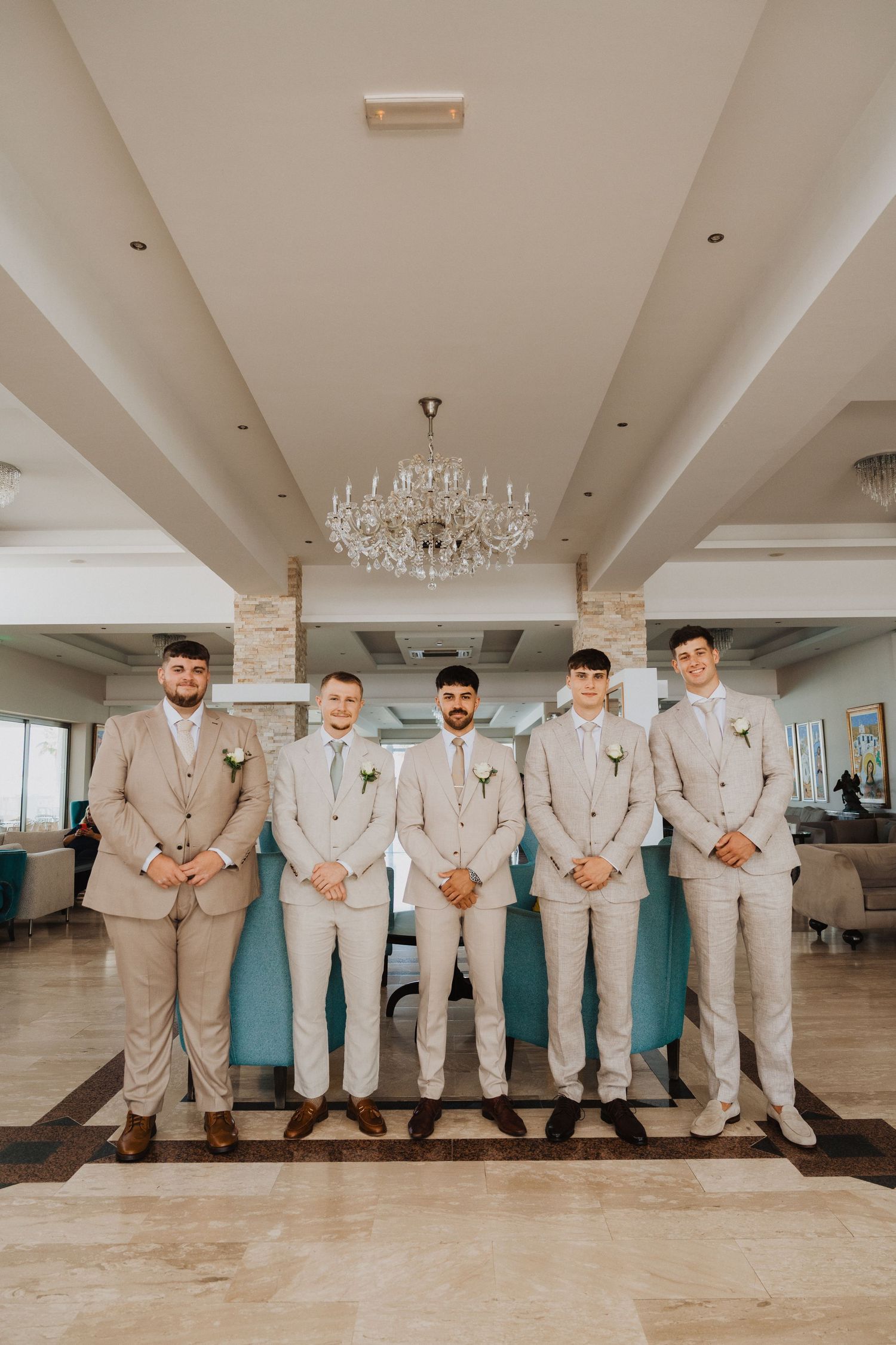 Wedding party in coordinated beige suits posing in elegant venue with chandelier.