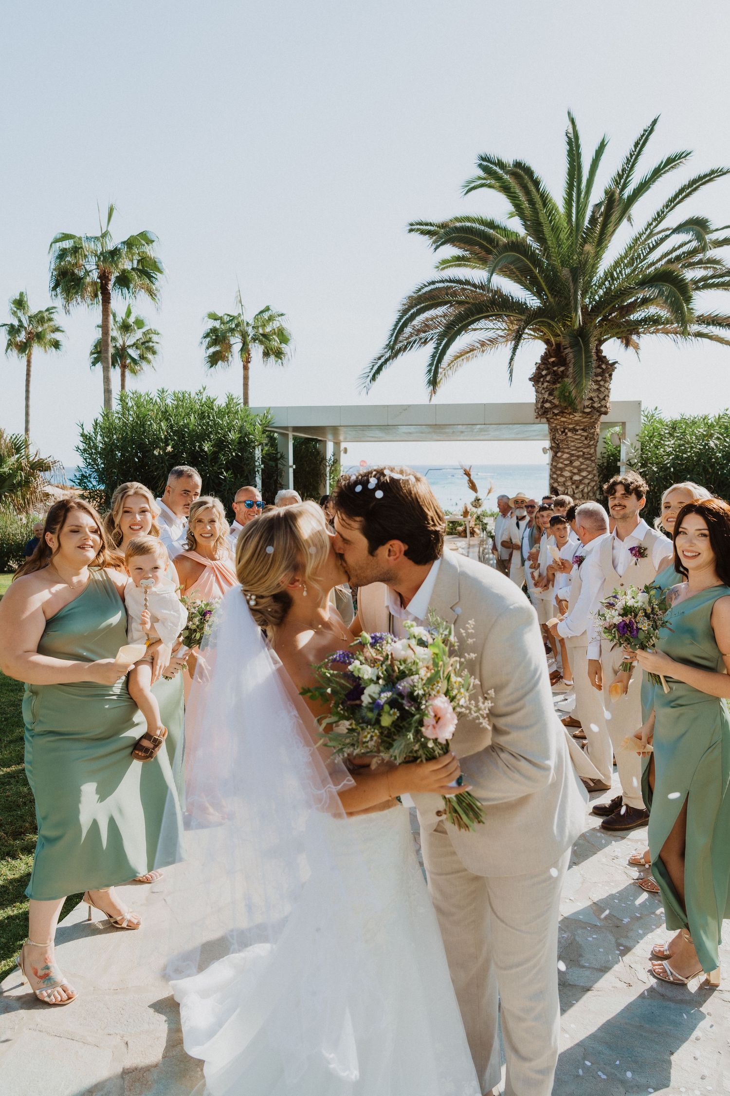 Wedding celebration with confetti toss along a palm-lined walkway under bright sunlight.