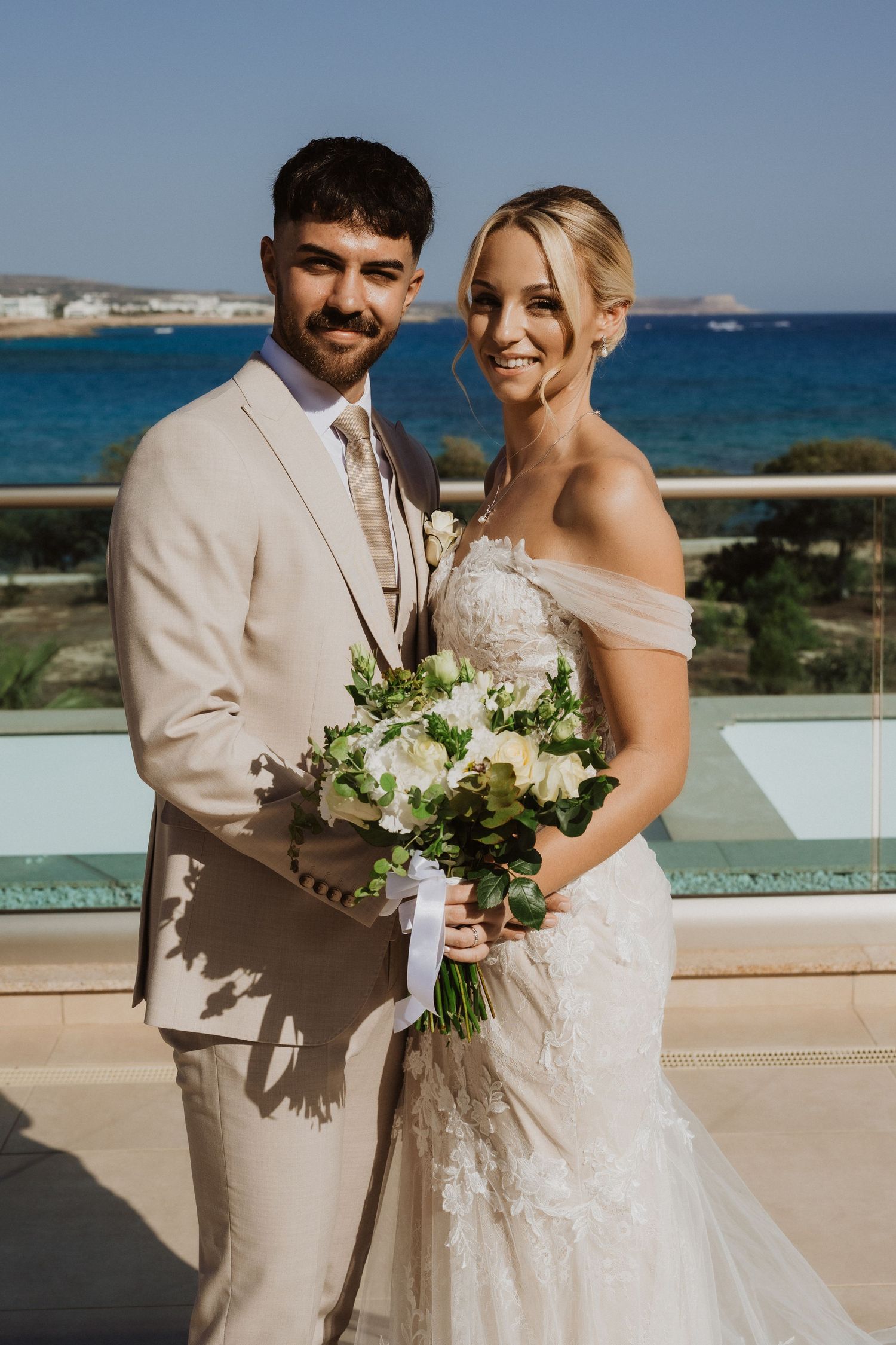 Bride and groom pose together on a seaside terrace with white floral bouquet.