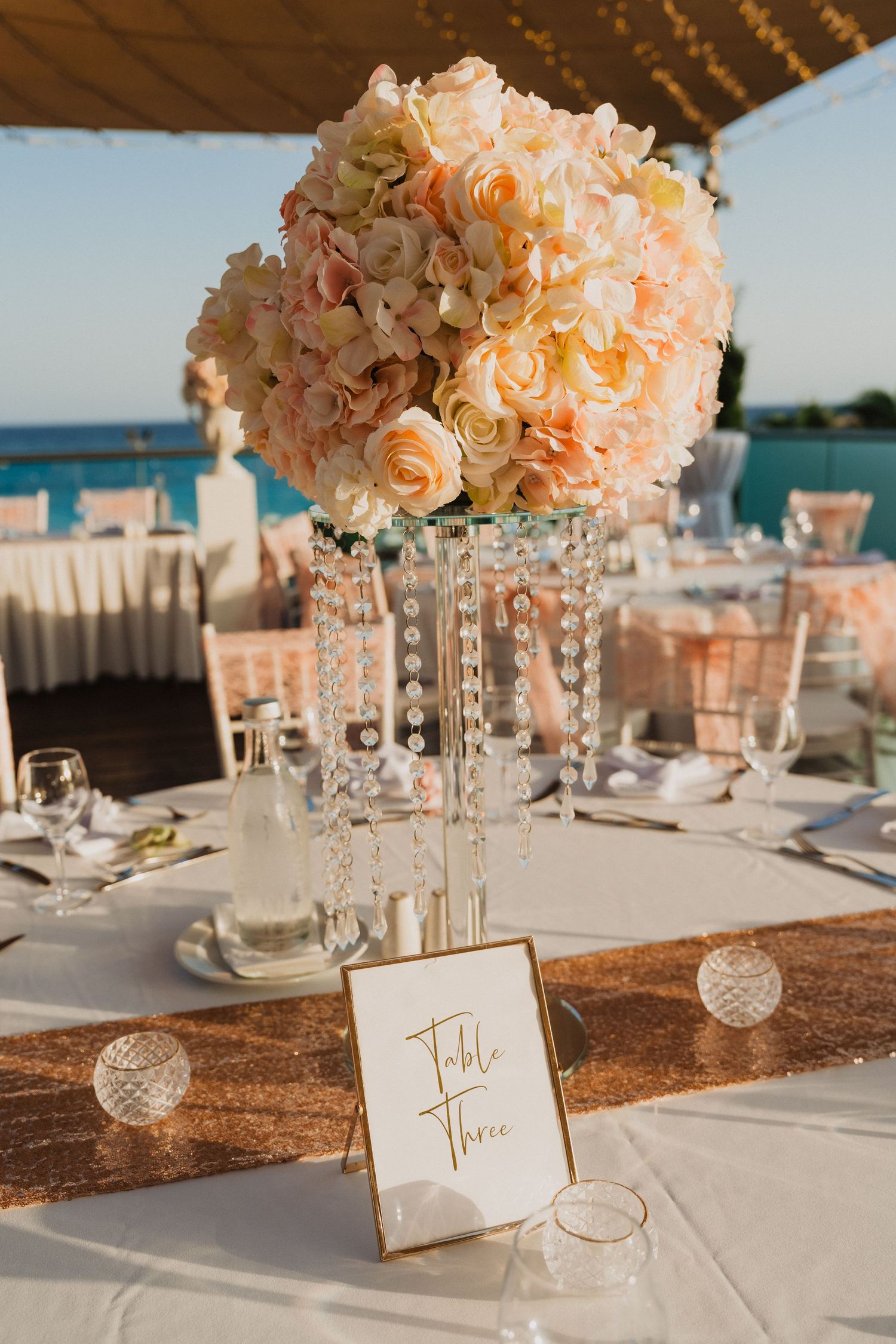 Elegant wedding table centerpiece with peach roses and crystal drops on a white tablecloth with gold sequin runner.