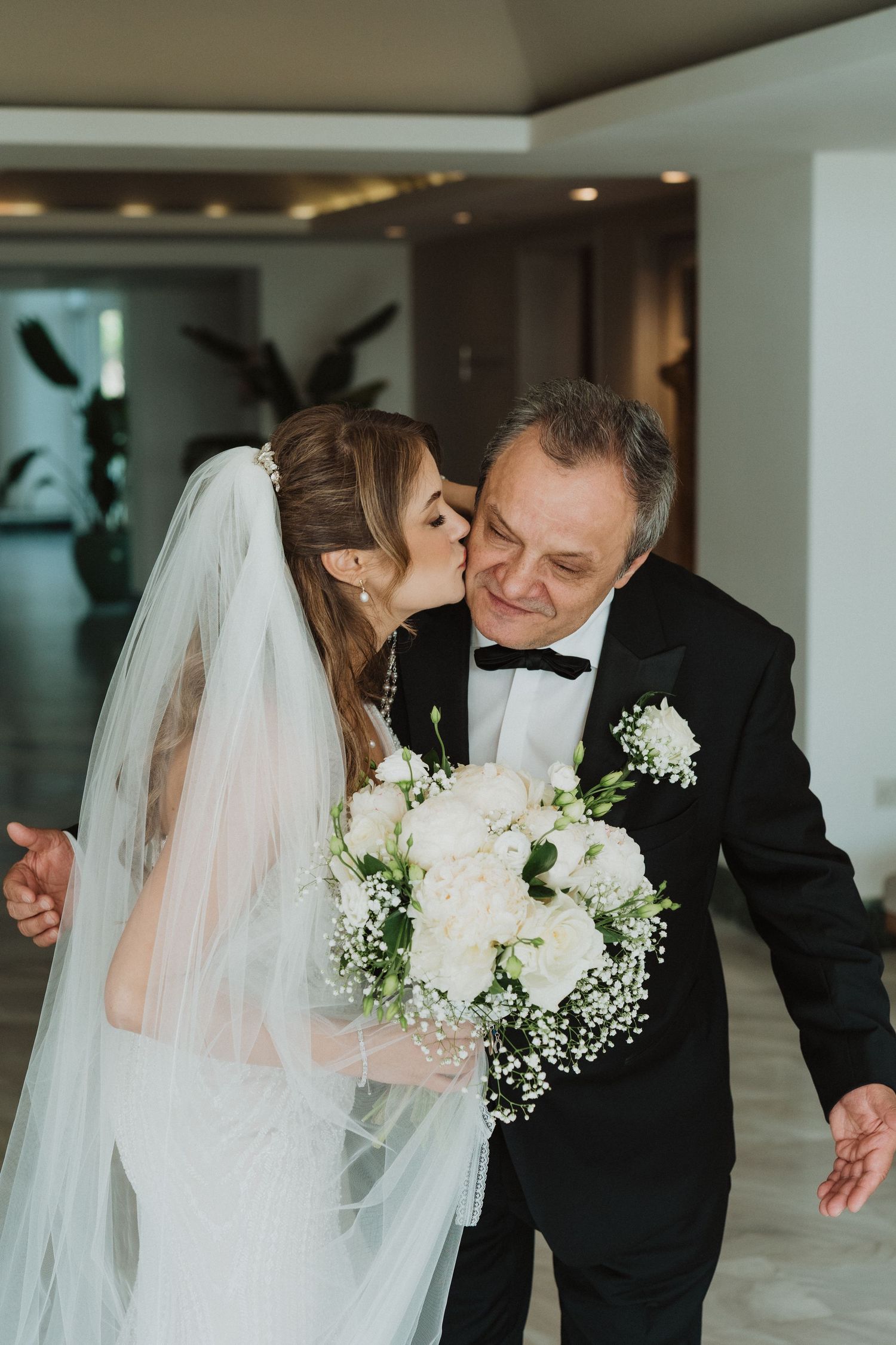 Bride in white gown kissing father on cheek while holding bouquet.