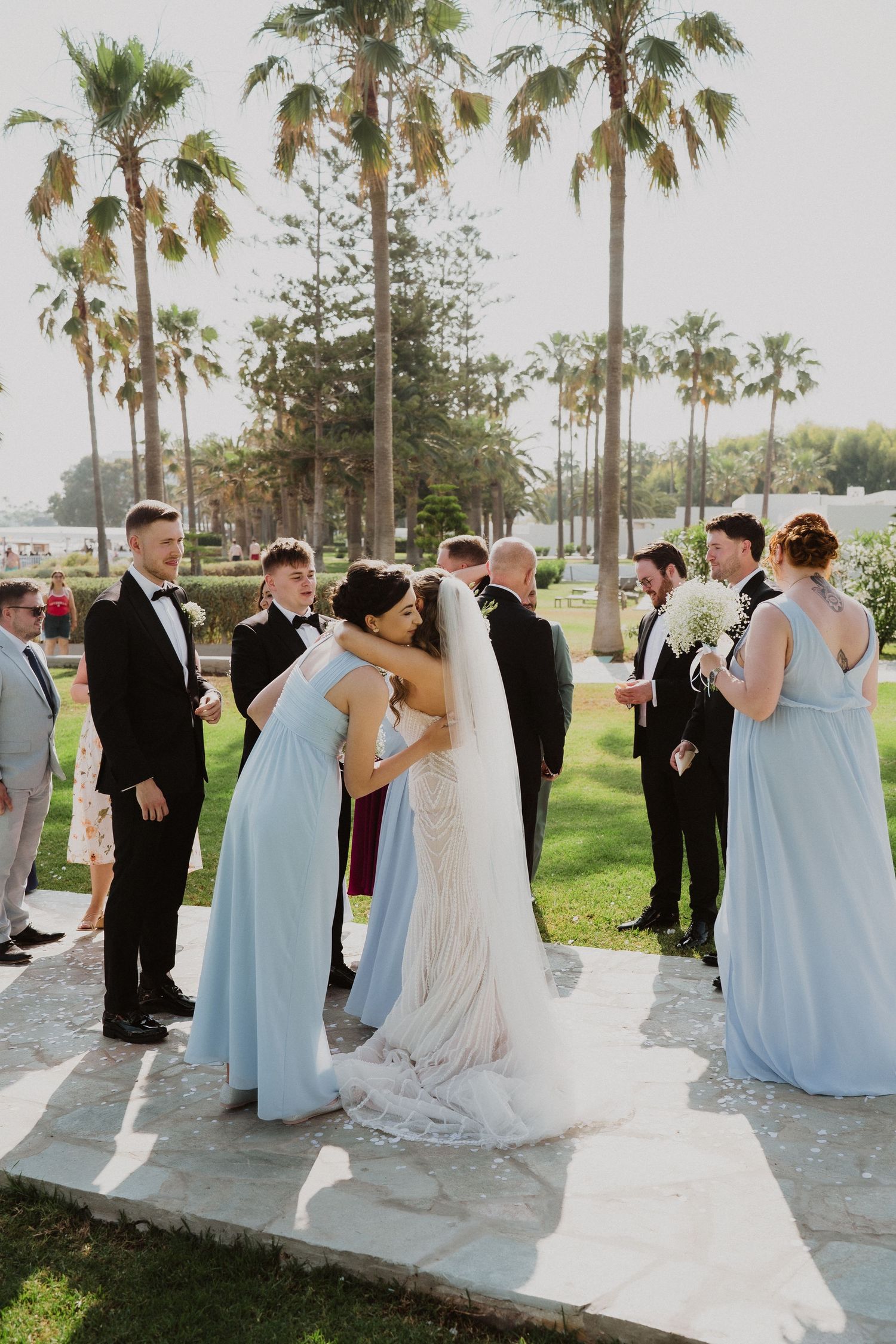 Wedding party greets bride in light blue bridesmaid dresses and black tuxedos among tall palm trees on a sunny day.