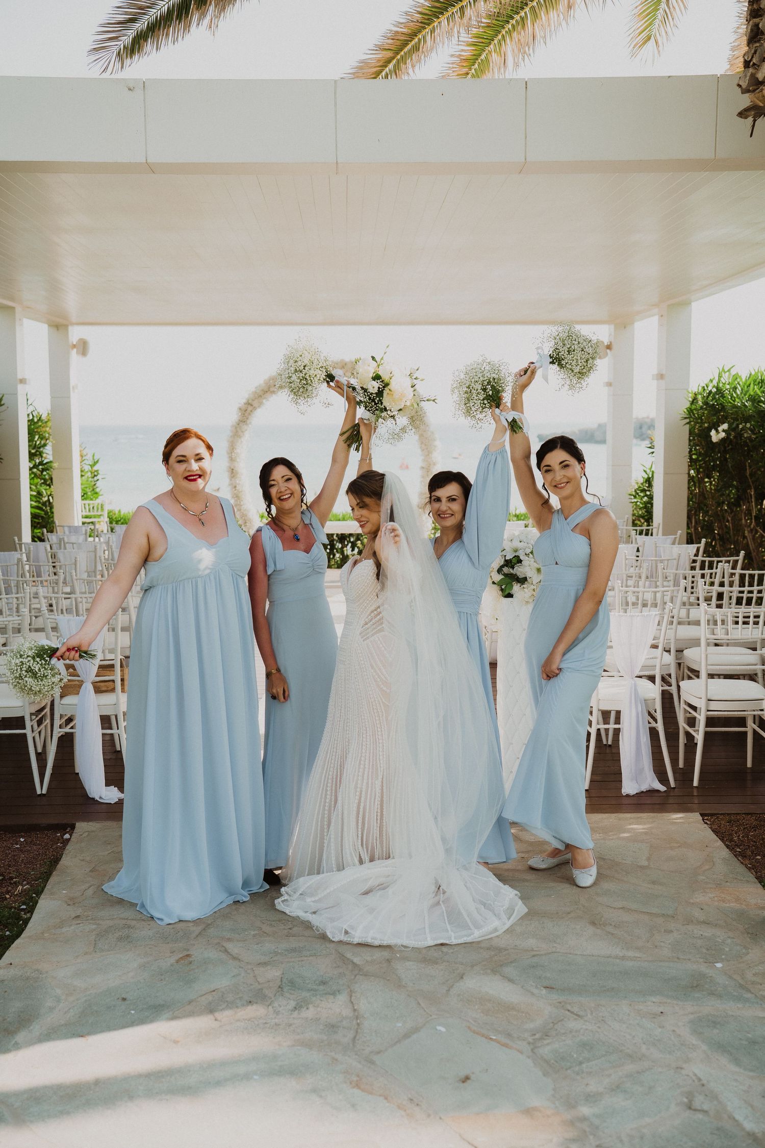 Bride celebrates with four bridesmaids in light blue dresses raising bouquets at outdoor wedding ceremony venue.